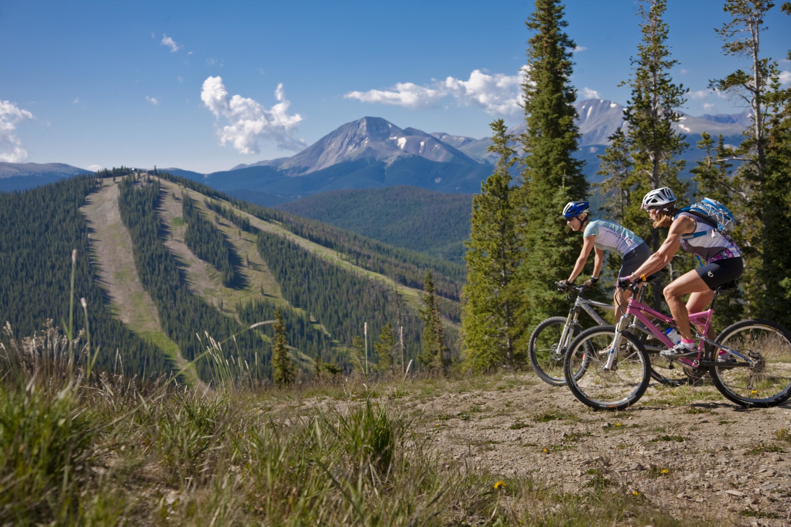 Two mountain bikers navigate a trail through a scenic landscape, featuring a backdrop of towering mountains and lush green forests. The sky is clear with a few clouds, and the sun is shining, highlighting the rugged terrain and the cyclists in action. Keystone Resort Bike Park mountain bike trail.