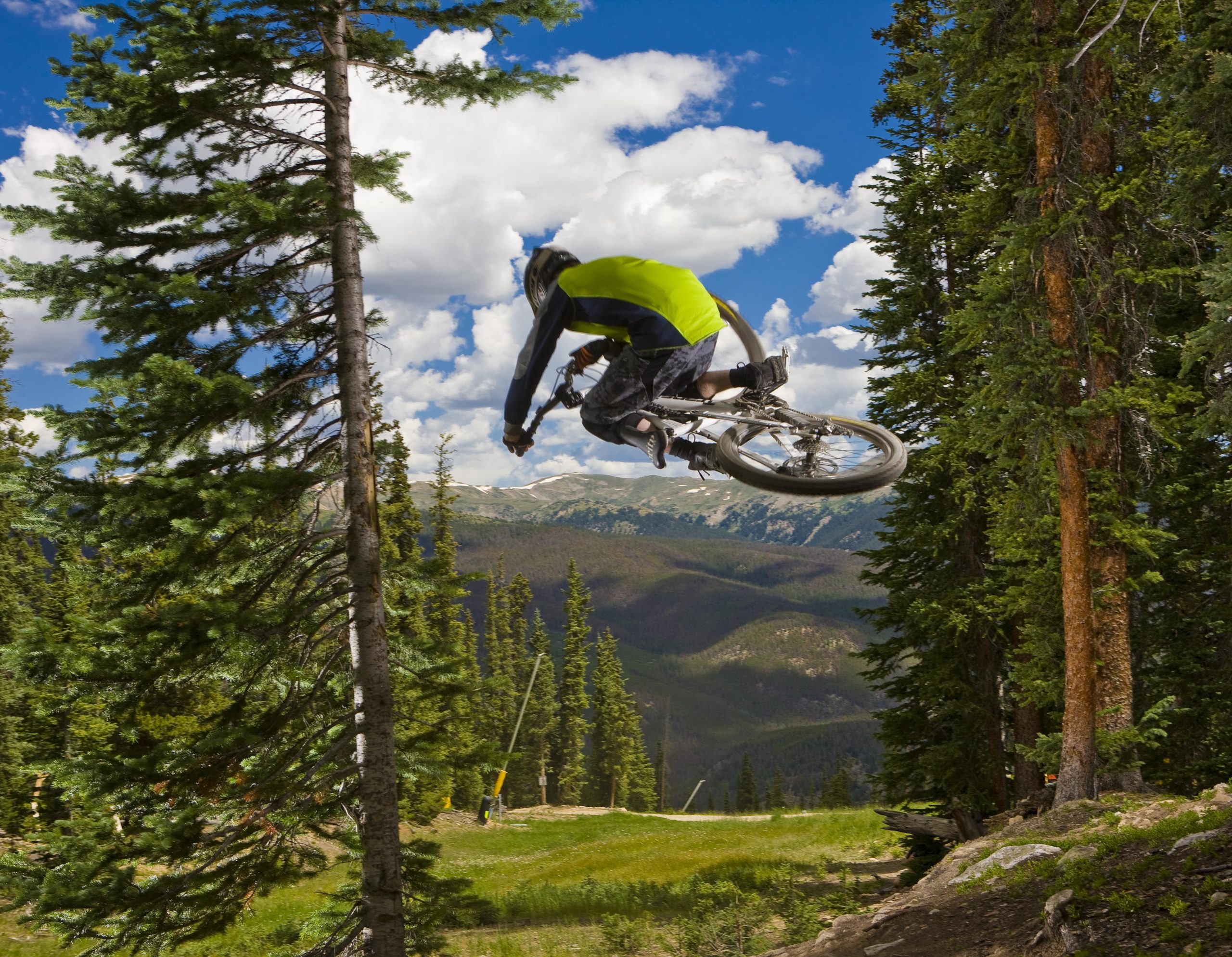 A mountain biker in a bright green jersey jumps off a dirt ramp, soaring through the air among tall pine trees. In the background, a scenic view of rolling hills and mountains can be seen under a partly cloudy blue sky. Keystone Resort Bike Park mountain bike trail.