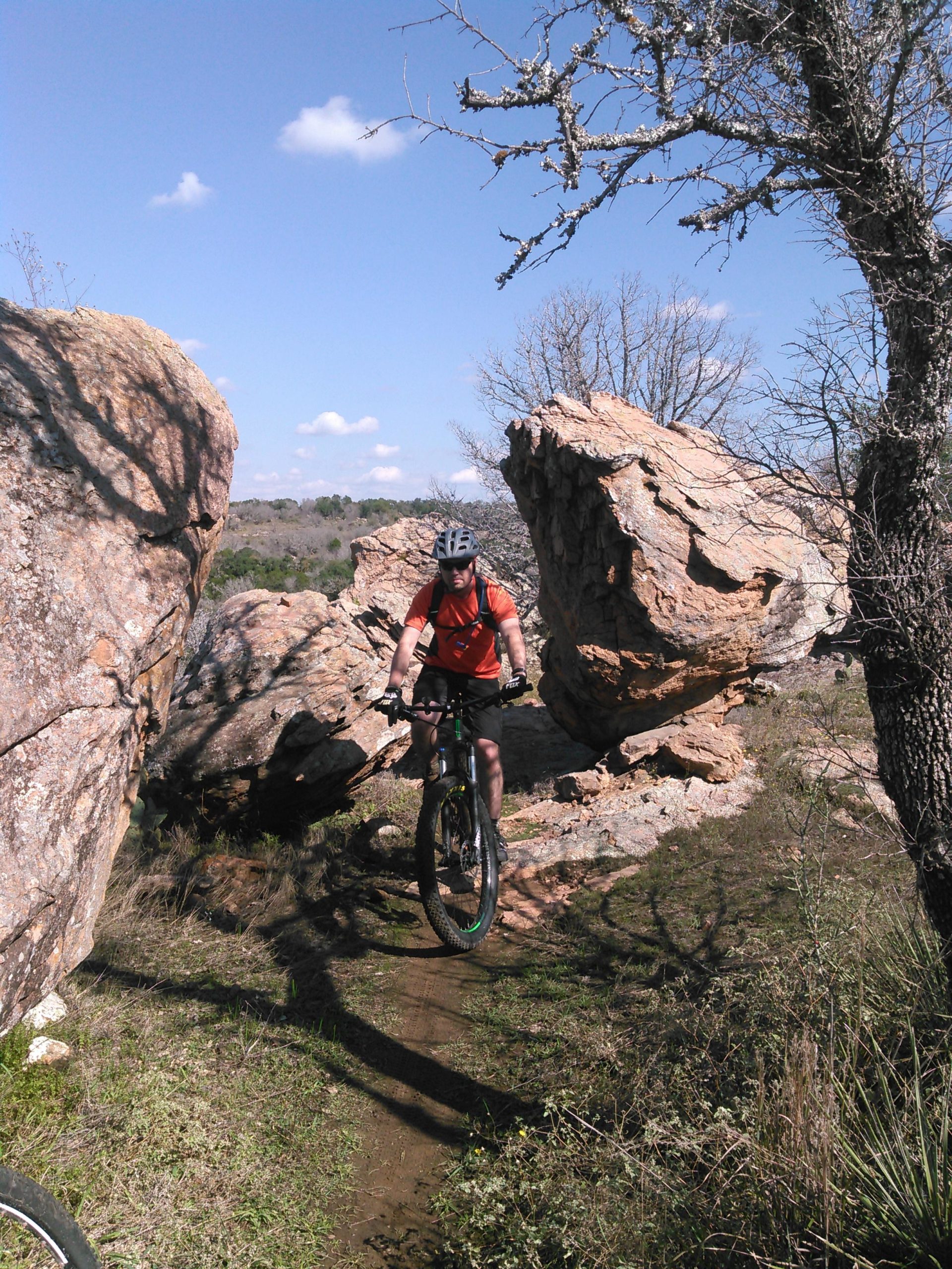 A mountain biker navigating a rocky trail surrounded by large boulders and sparse vegetation under a clear blue sky. Reveille Peak Ranch mountain bike trail.