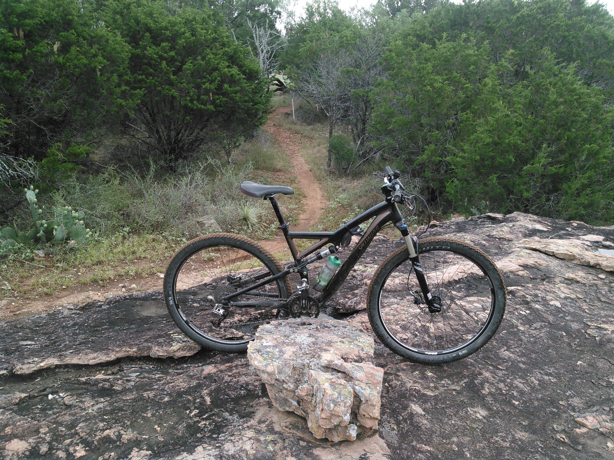 A black mountain bike resting on a rock, with a dirt trail winding through green vegetation in the background. Trees and cacti are visible, indicating an outdoor, rugged environment. Reveille Peak Ranch mountain bike trail.