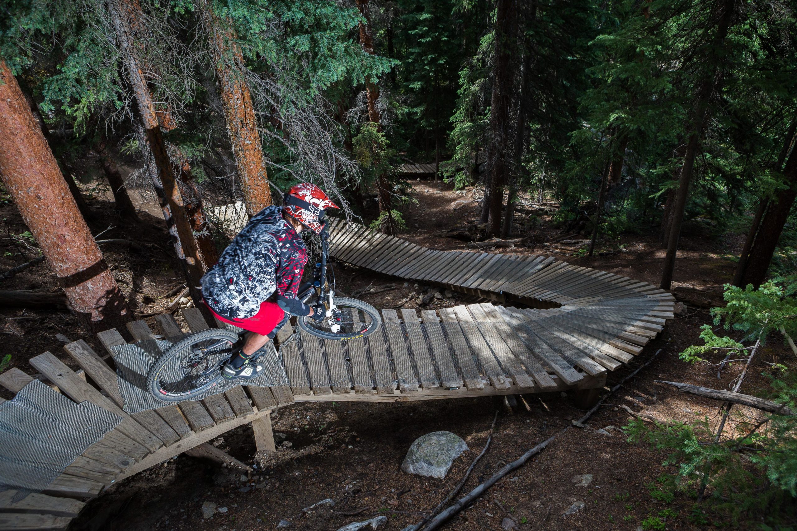 A mountain biker rides on a wooden boardwalk trail winding through a dense forest of tall trees. The cyclist, wearing a helmet and colorful gear, is navigating a curved section of the path, showcasing a dynamic downhill position. Sunlight filters through the trees, highlighting the natural setting and the intricate design of the trail. Keystone Resort Bike Park mountain bike trail.