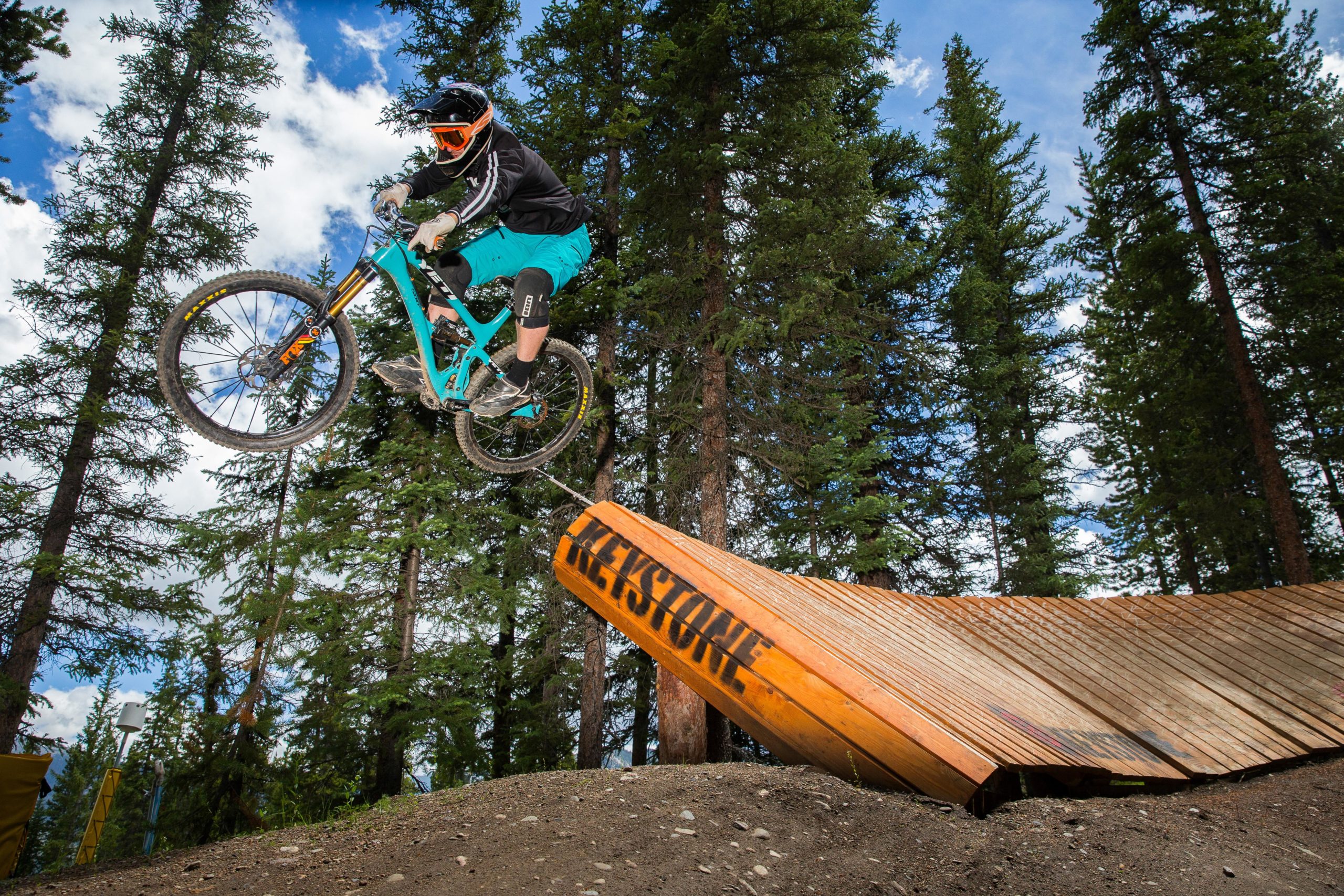 A mountain biker in a black helmet and orange goggles performs a jump off a wooden ramp in a forested area. The biker is airborne above the ramp, surrounded by tall evergreen trees and a bright blue sky with scattered clouds. Keystone Resort Bike Park mountain bike trail.