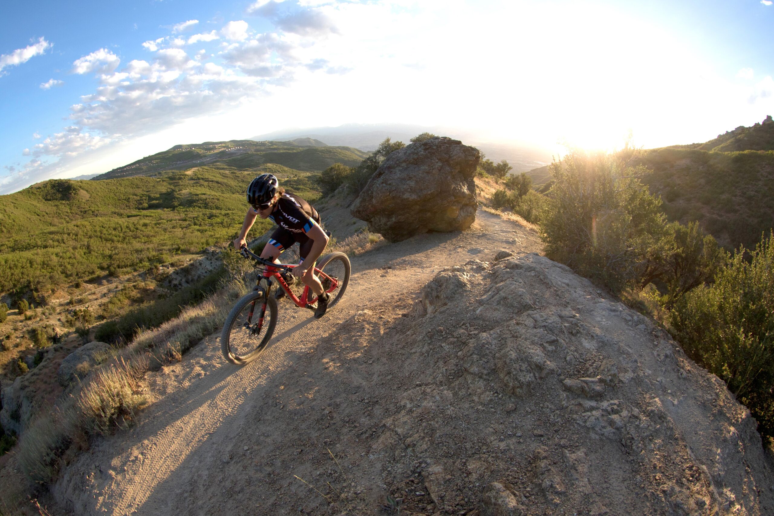 Pivot LES 27.5: A mountain biker rides along a dirt trail with rocky terrain, surrounded by lush green hills under a partly cloudy sky. The sun is low in the sky, casting a warm glow over the landscape.