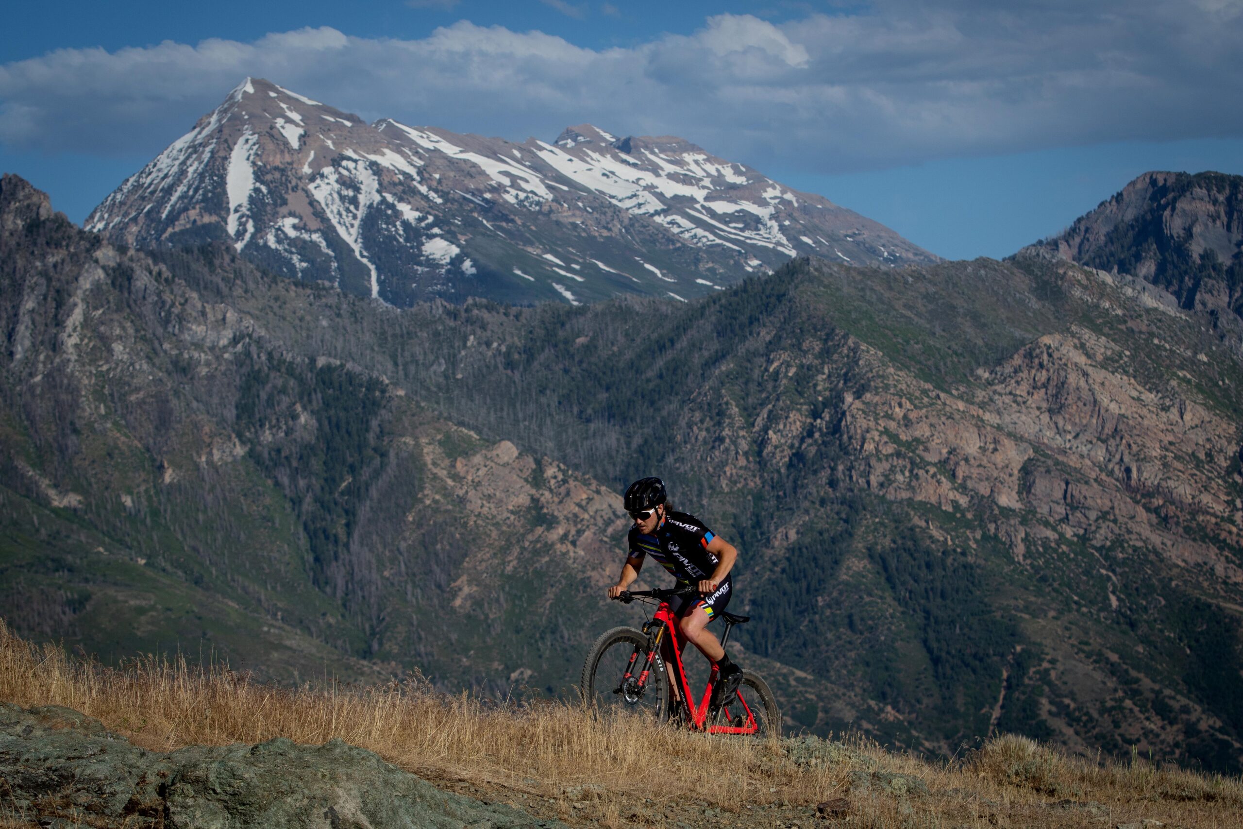 Pivot LES 27.5: A mountain biker navigating a rocky trail with a red bicycle, set against a backdrop of snow-capped mountains and blue skies. The lush greenery and rugged terrain are visible in the surrounding landscape, highlighting the beauty of outdoor biking adventures.