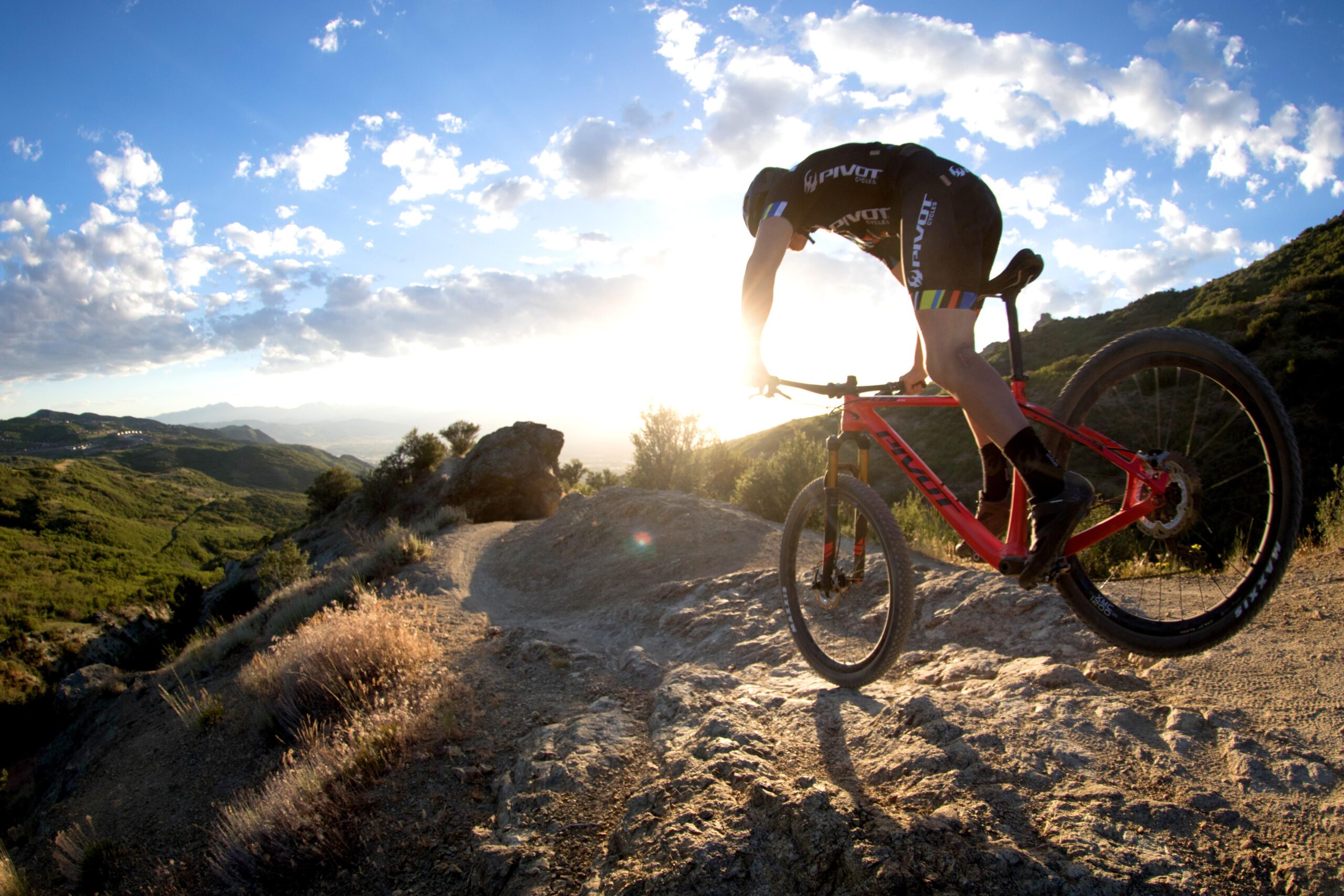 Pivot LES 27.5: A cyclist performing a jump on a mountain bike, soaring over a rocky trail with a scenic landscape of green hills and a bright sky in the background. The sun sets behind the hills, casting a warm glow on the surroundings.