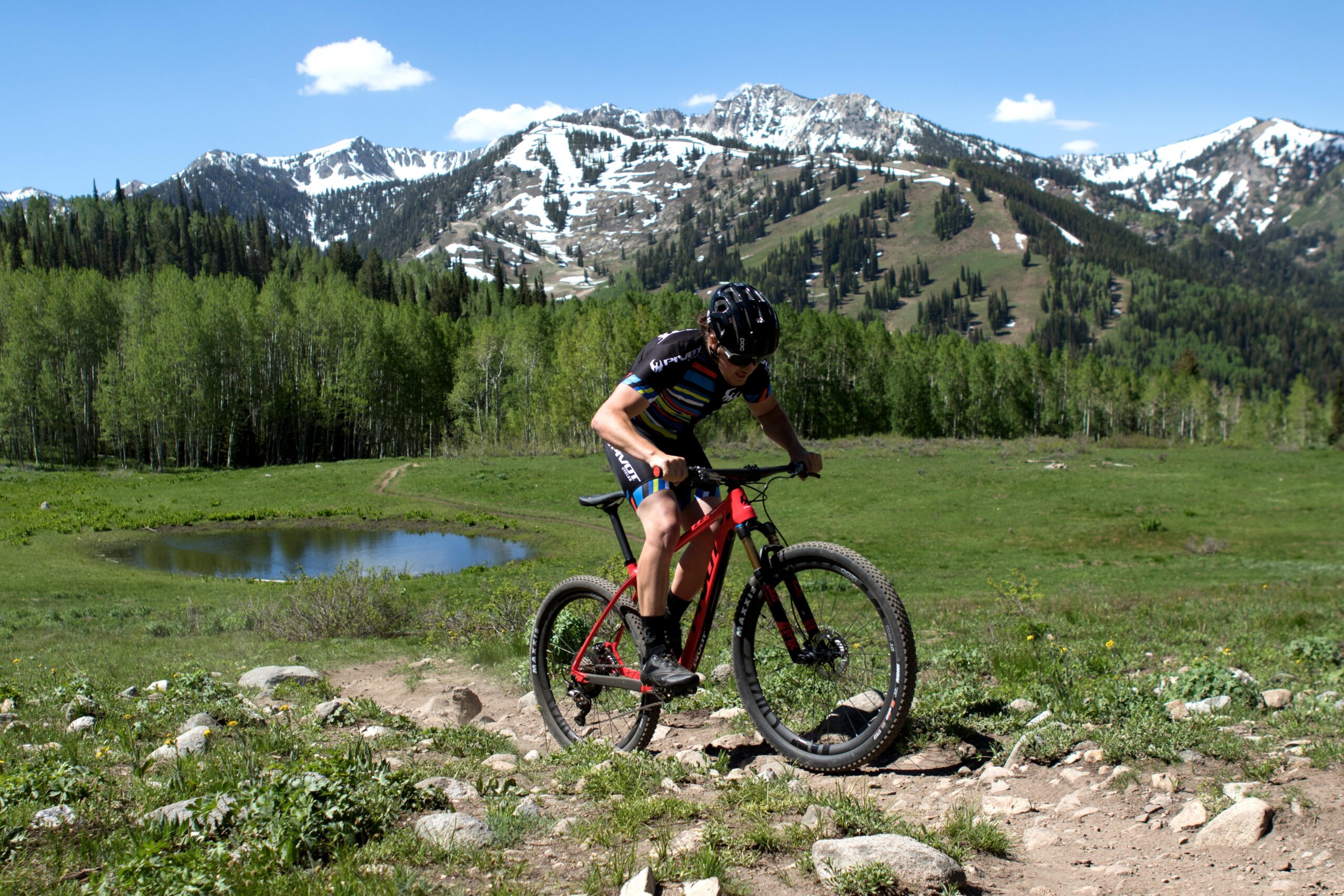 Pivot LES 27.5: A mountain biker riding over rocky terrain in a scenic landscape with green grass, a small pond, and snow-capped mountains in the background under a clear blue sky.