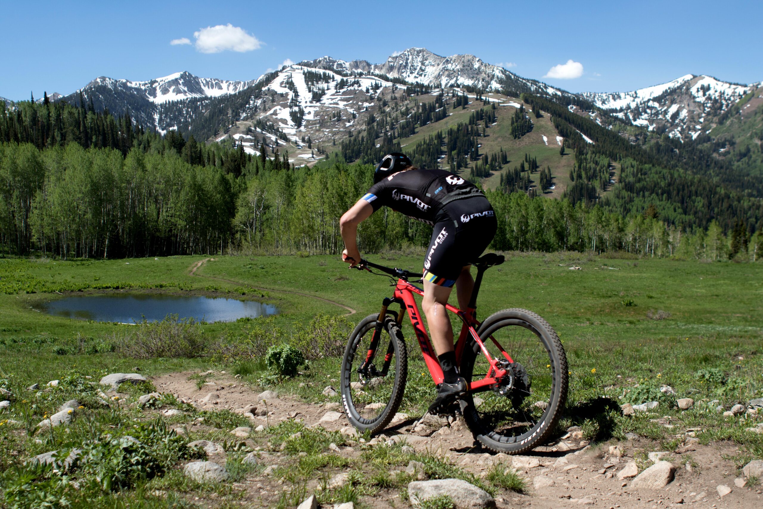 Pivot LES 27.5: A mountain biker navigates a rocky trail in a lush green landscape, with a small pond nearby and snow-capped mountains in the background under a clear blue sky.