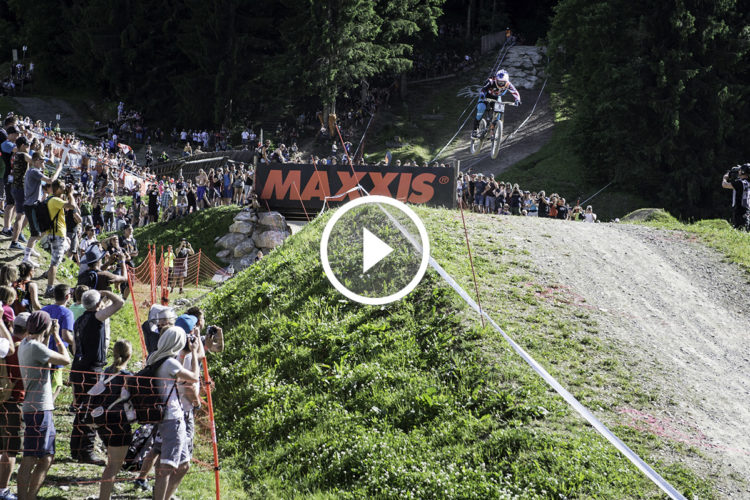 A mountain biker leaps off a dirt ramp during a downhill race, with a crowd of spectators and photographers watching from the side. The scene features lush greenery and a prominent advertising banner for Maxxis in the background.