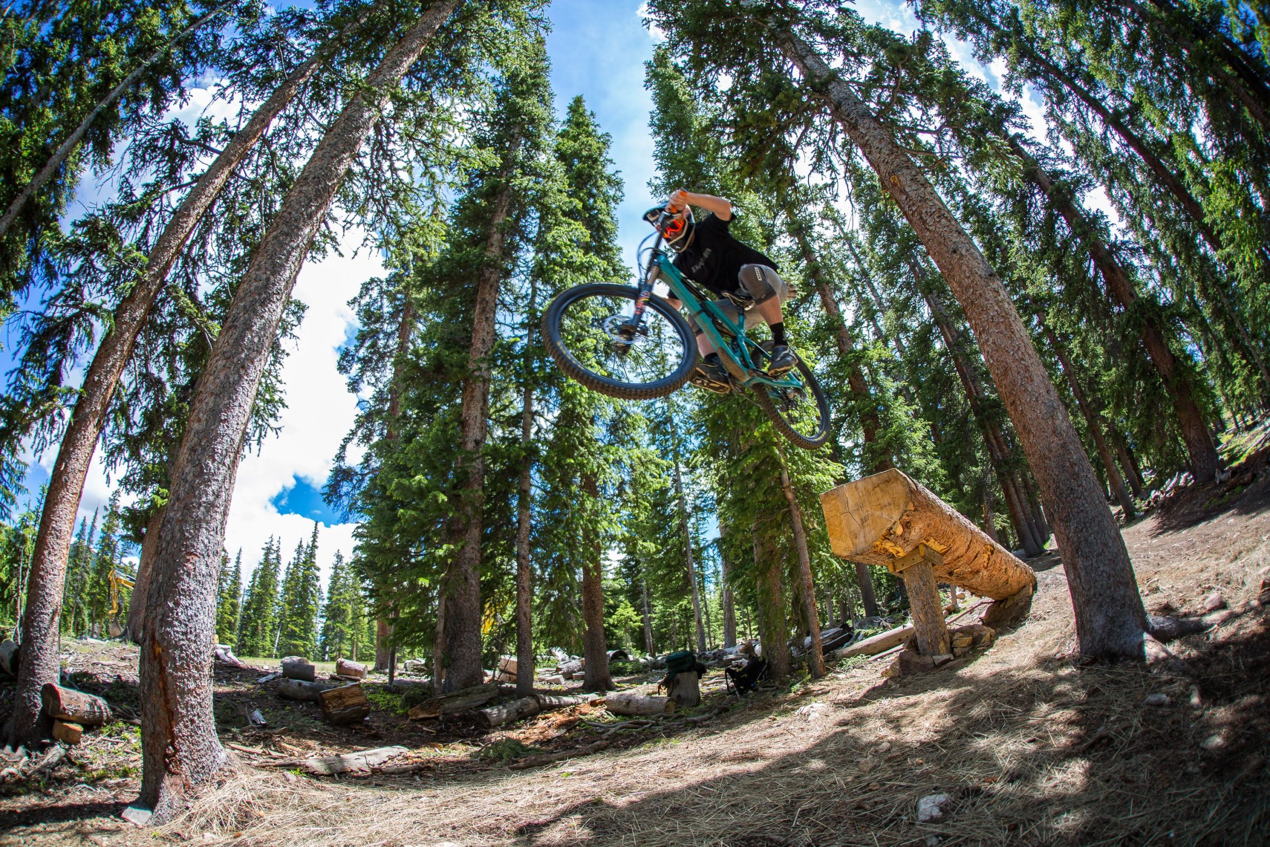 A mountain biker mid-air, performing a jump over a wooden log, surrounded by tall trees in a forest. The sky is clear with a few clouds visible. Keystone Resort Bike Park mountain bike trail.