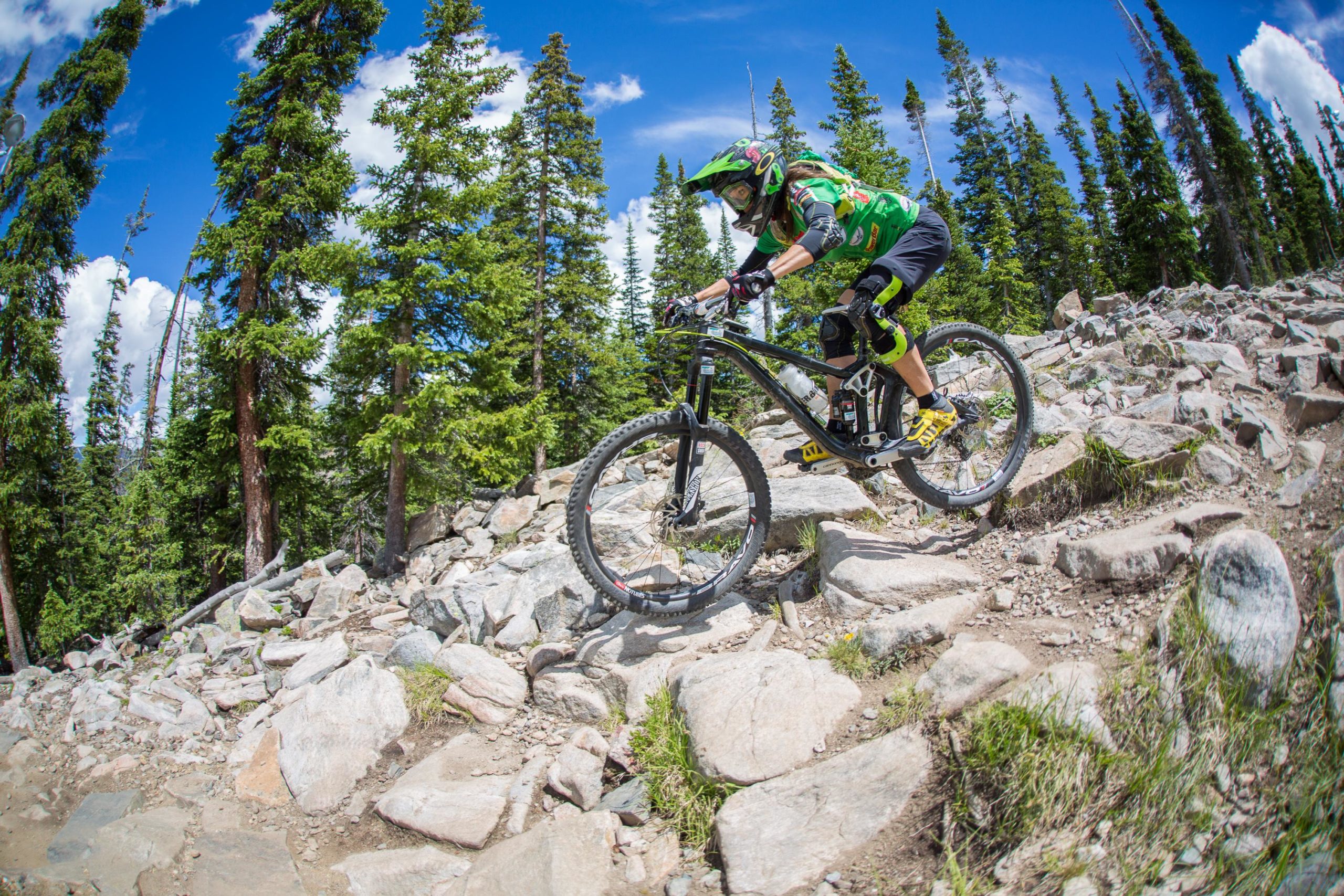 A mountain biker rides down a rocky trail surrounded by tall green trees, showcasing a dynamic moment of the sport against a bright blue sky with fluffy white clouds. The rider is wearing protective gear and a helmet, and the bicycle features a sturdy frame designed for rugged terrain. Keystone Resort Bike Park mountain bike trail.