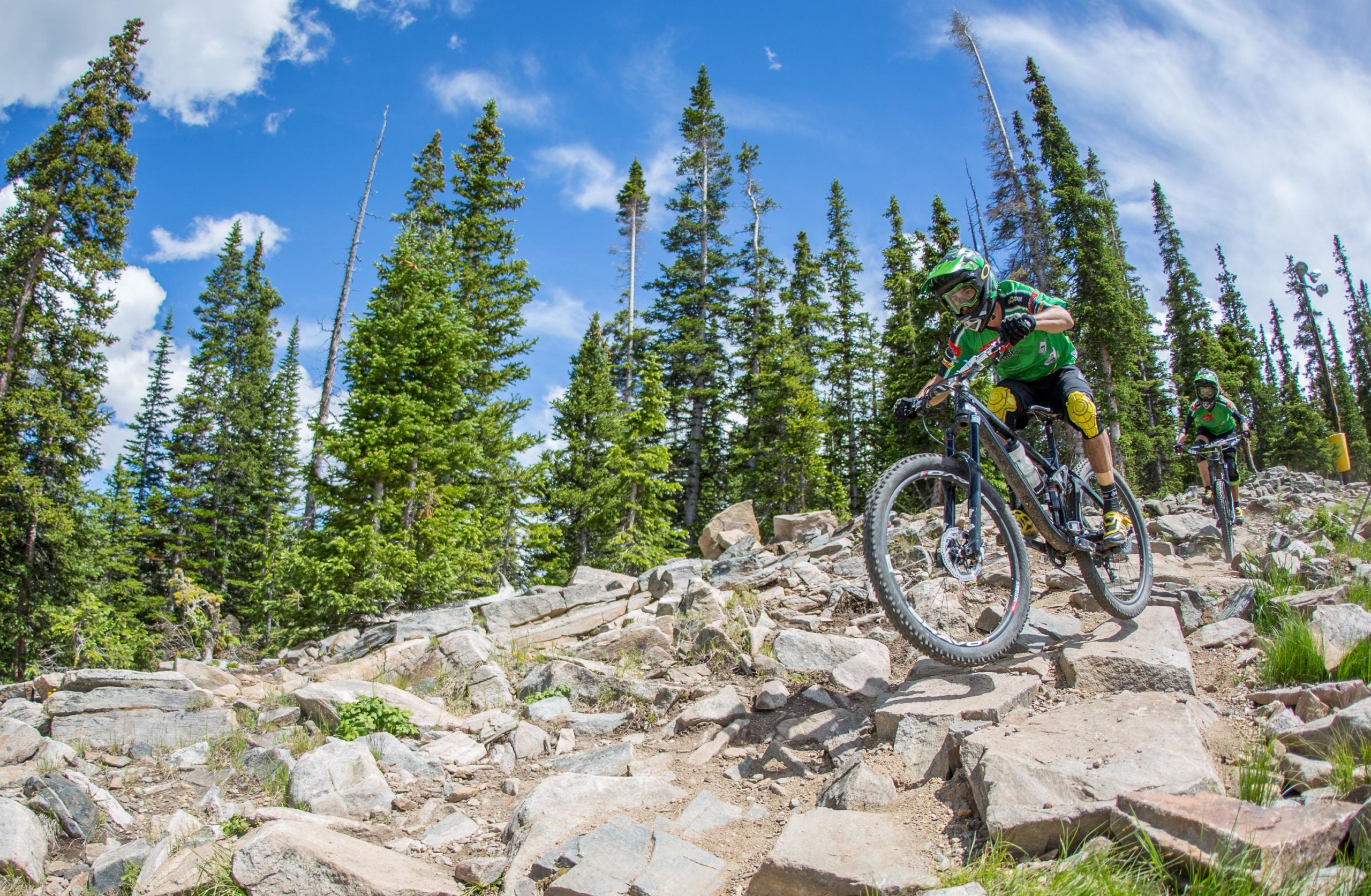 A mountain biker navigating a rocky trail through a lush forest, with tall pine trees and a blue sky in the background. Another biker follows closely behind. Keystone Resort Bike Park mountain bike trail.