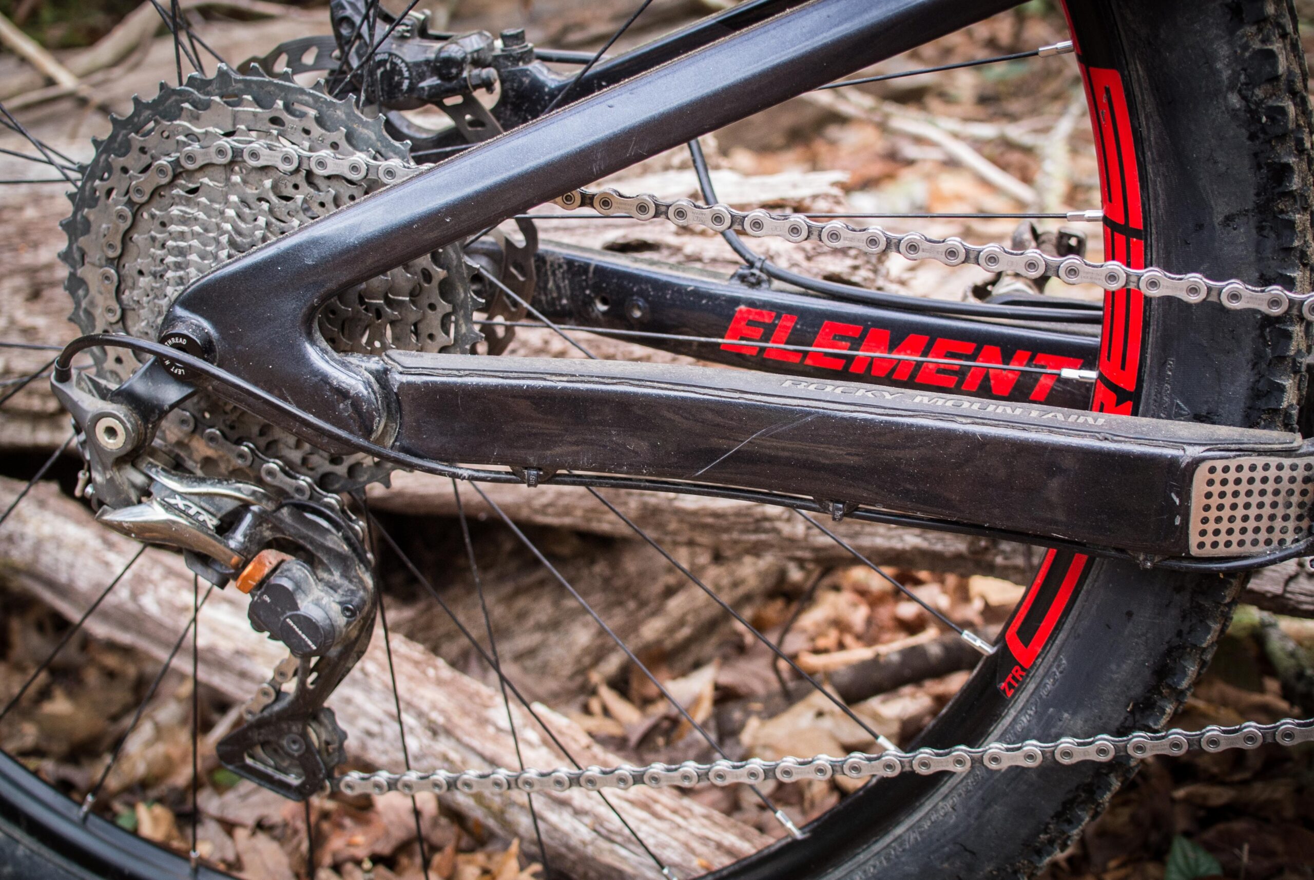 Rocky Mountain Element: Close-up view of a mountain bike's drivetrain, featuring a chain, derailleur, and cassette. The frame displays the "Element" logo, set against a backdrop of wooden logs and fallen leaves. The components show signs of use, highlighting a rugged outdoor environment.