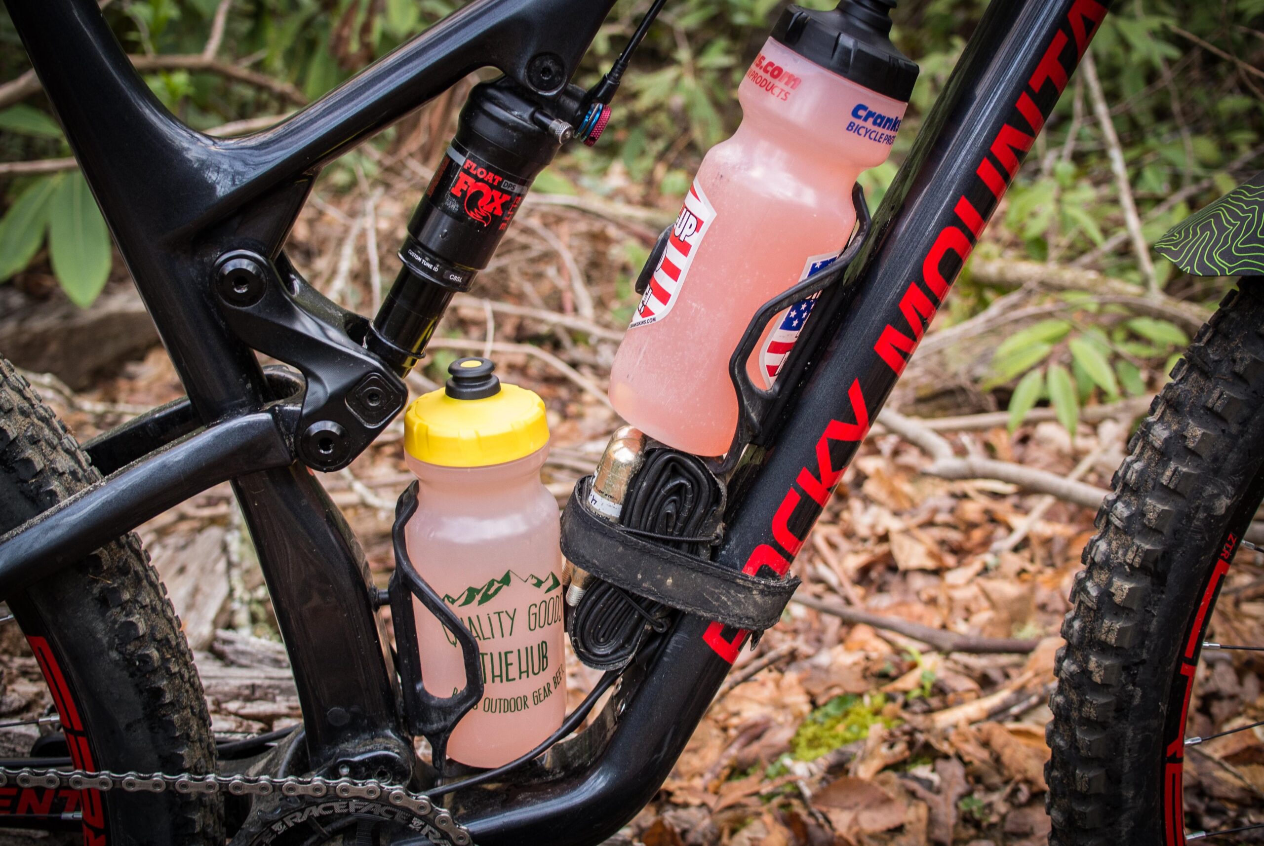 Rocky Mountain Element: Alt text: A close-up of a black mountain bike frame featuring two water bottles in the frame's bottle holders. One bottle has a yellow lid and a printed label, while the other has an American flag design. The bike's suspension and chain are also visible, set against a backdrop of leaves and greenery.