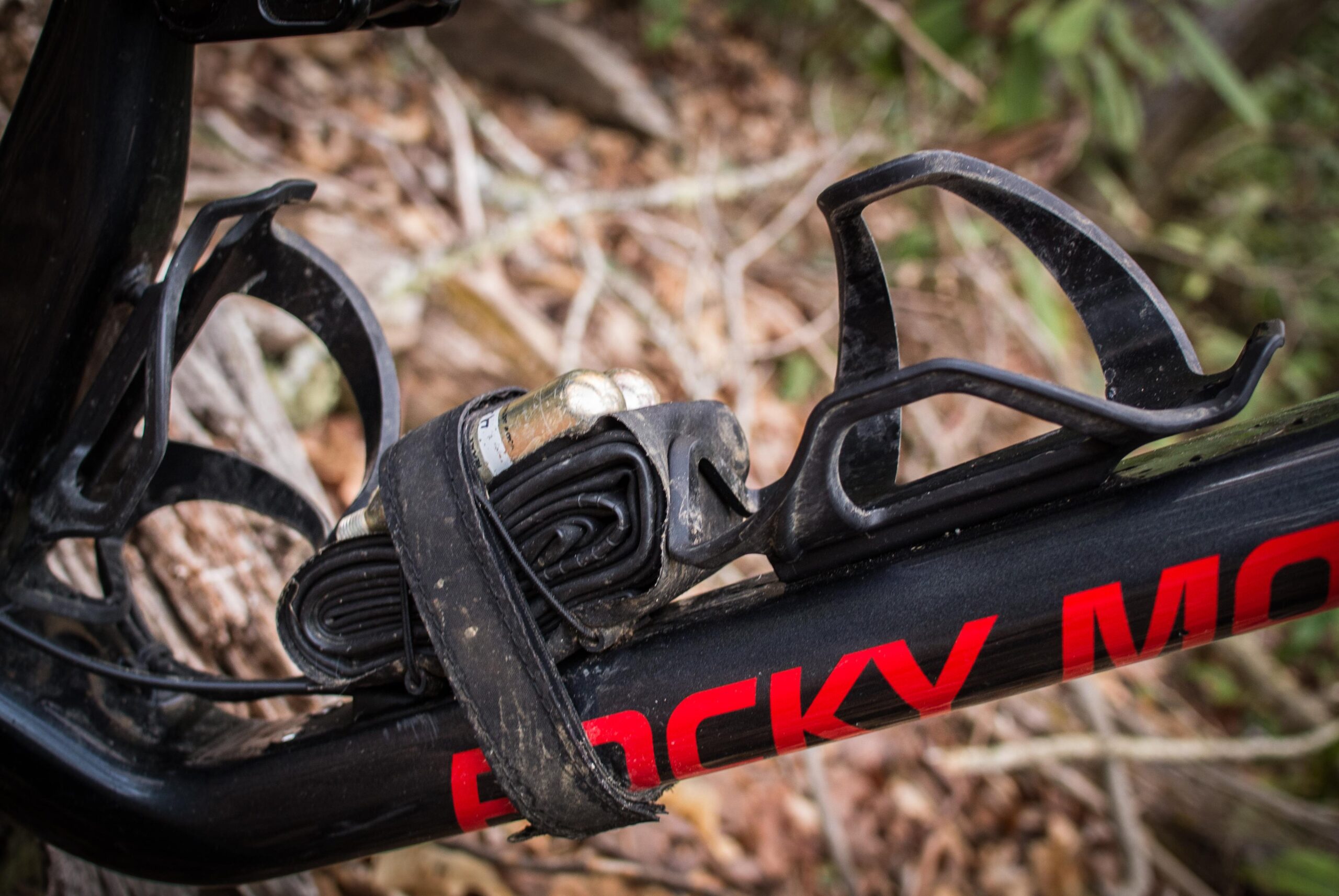 Rocky Mountain Element: A close-up of a mountain bike frame with a black water bottle cage holding a rolled-up inner tube secured with a strap. The bike's body features a red logo and is set against a background of earthy leaves and foliage.