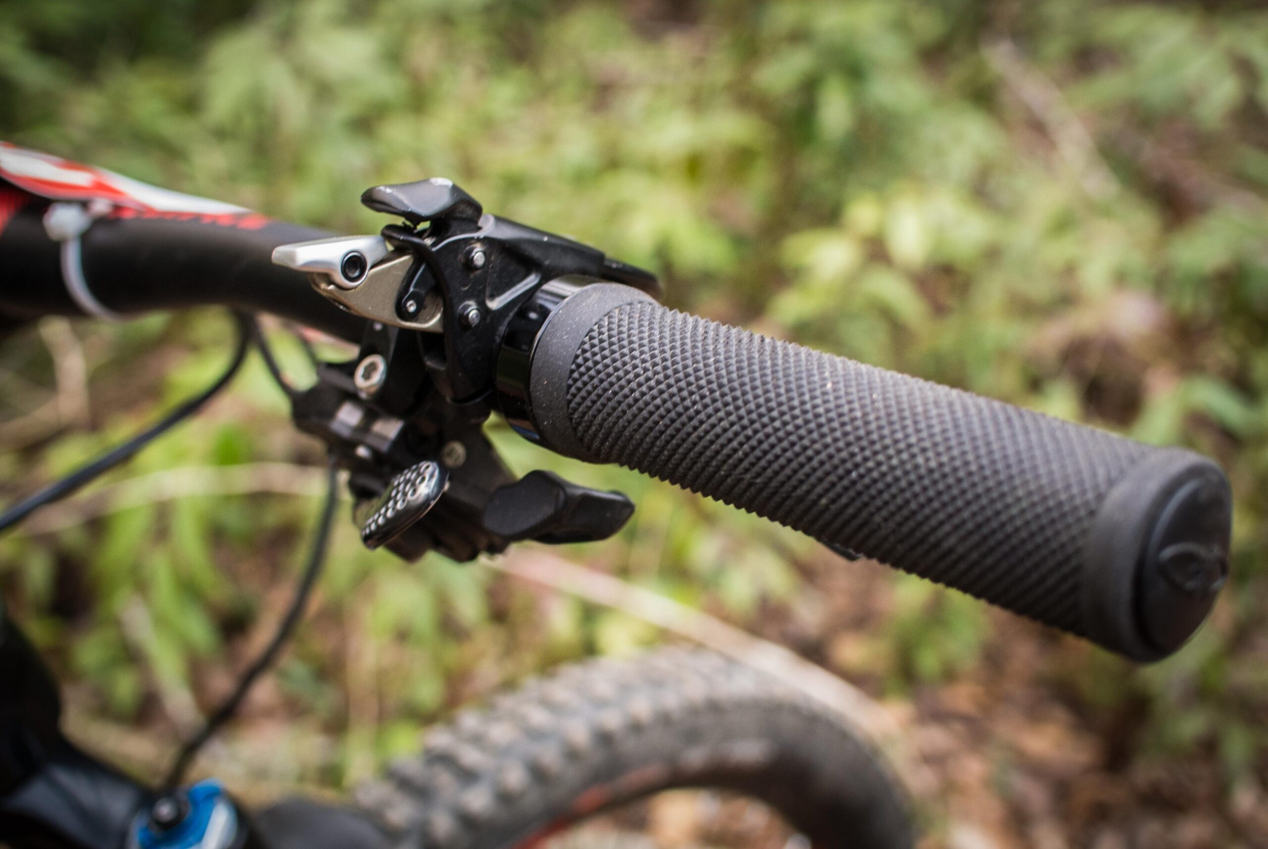 Rocky Mountain Element: Close-up of a mountain bike handlebar featuring a textured black grip, gear shifters, and brake lever against a natural, blurred green backdrop.
