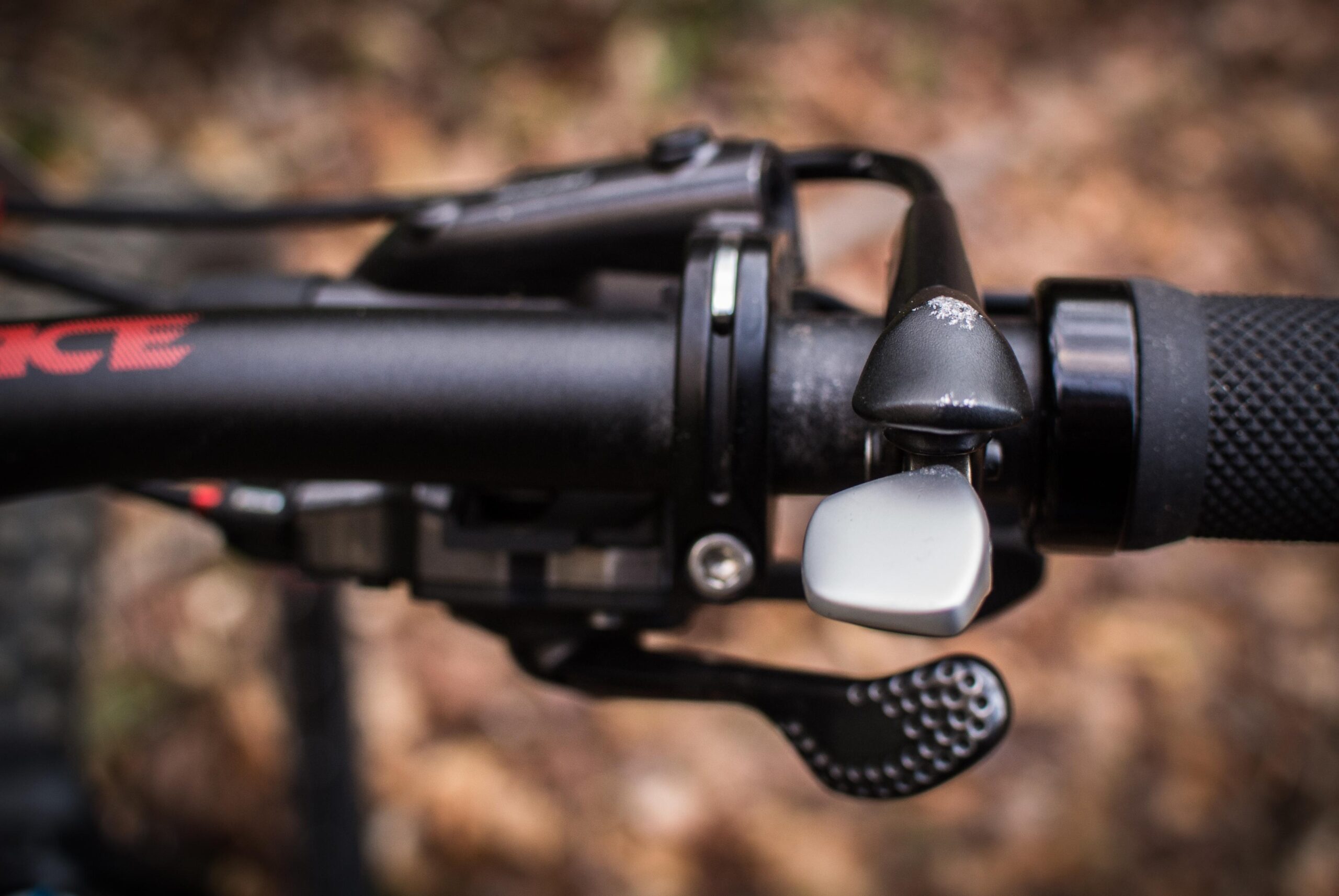 Rocky Mountain Element: Close-up view of a mountain bike handlebar featuring a gear shift lever, brake lever, and textured grip, with a blurred background of autumn foliage.