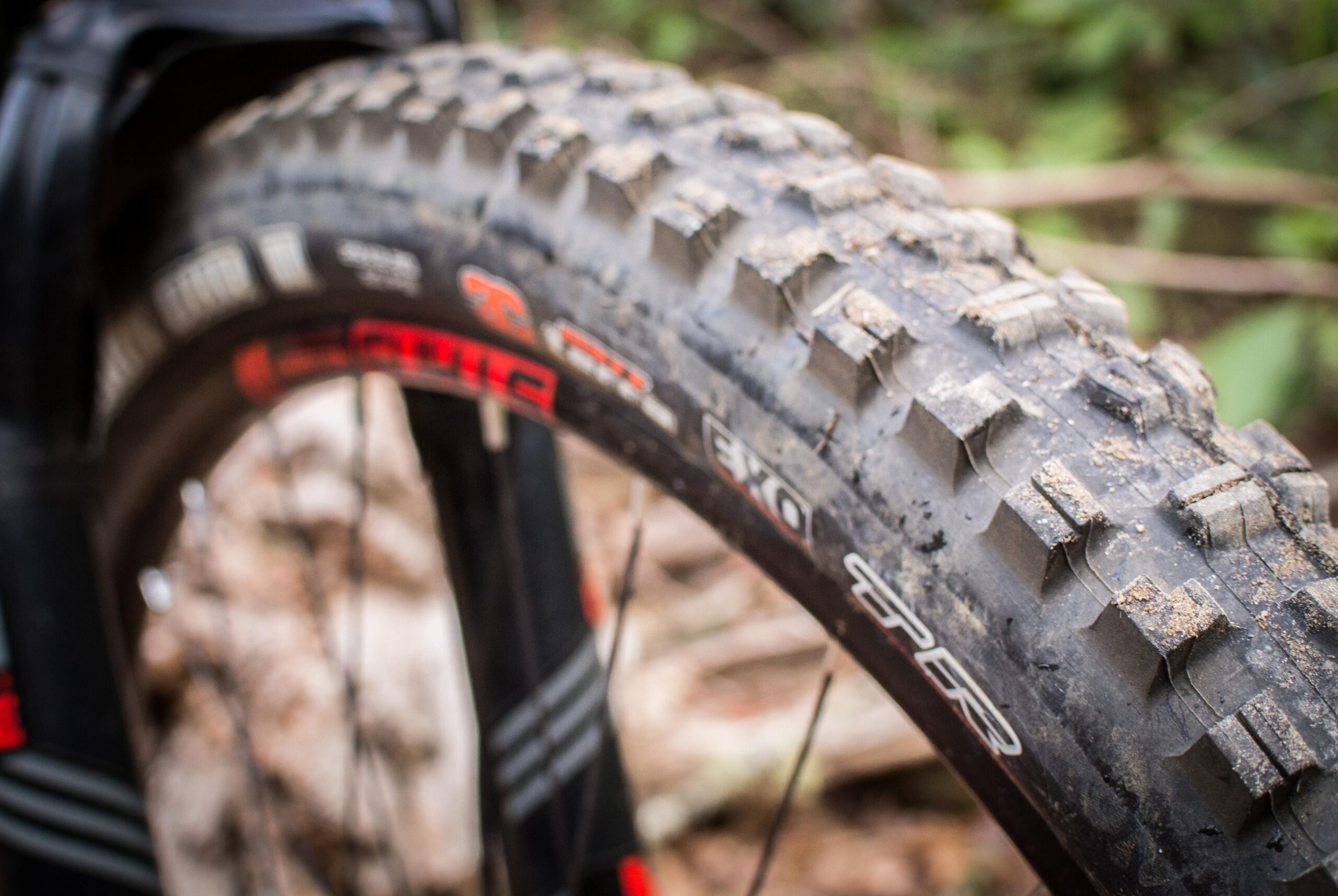 Rocky Mountain Element: Close-up of a mountain bike tire with a knobby tread design, showing dirt and debris, set against a blurred background of natural foliage.