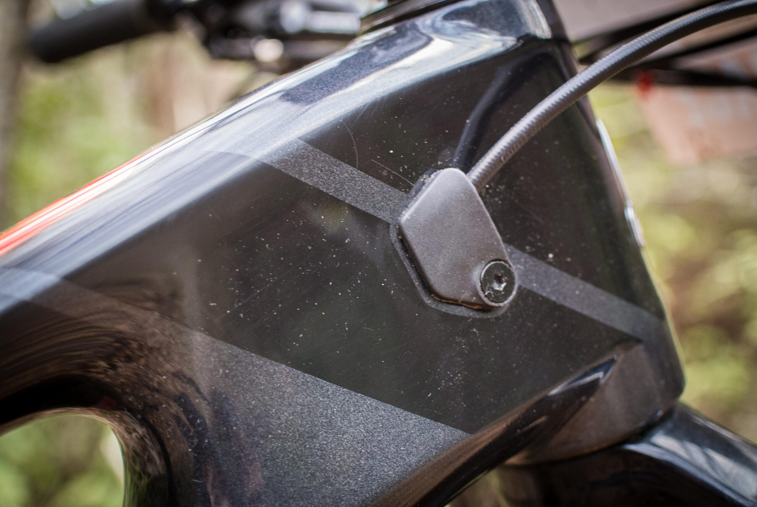 Rocky Mountain Element: Close-up of the frame of a bike, featuring a sleek dark finish with a textured pattern and visible scratches. A small black cable guide is attached to the frame, with a screw visible. The background consists of blurred greenery, suggesting an outdoor setting.
