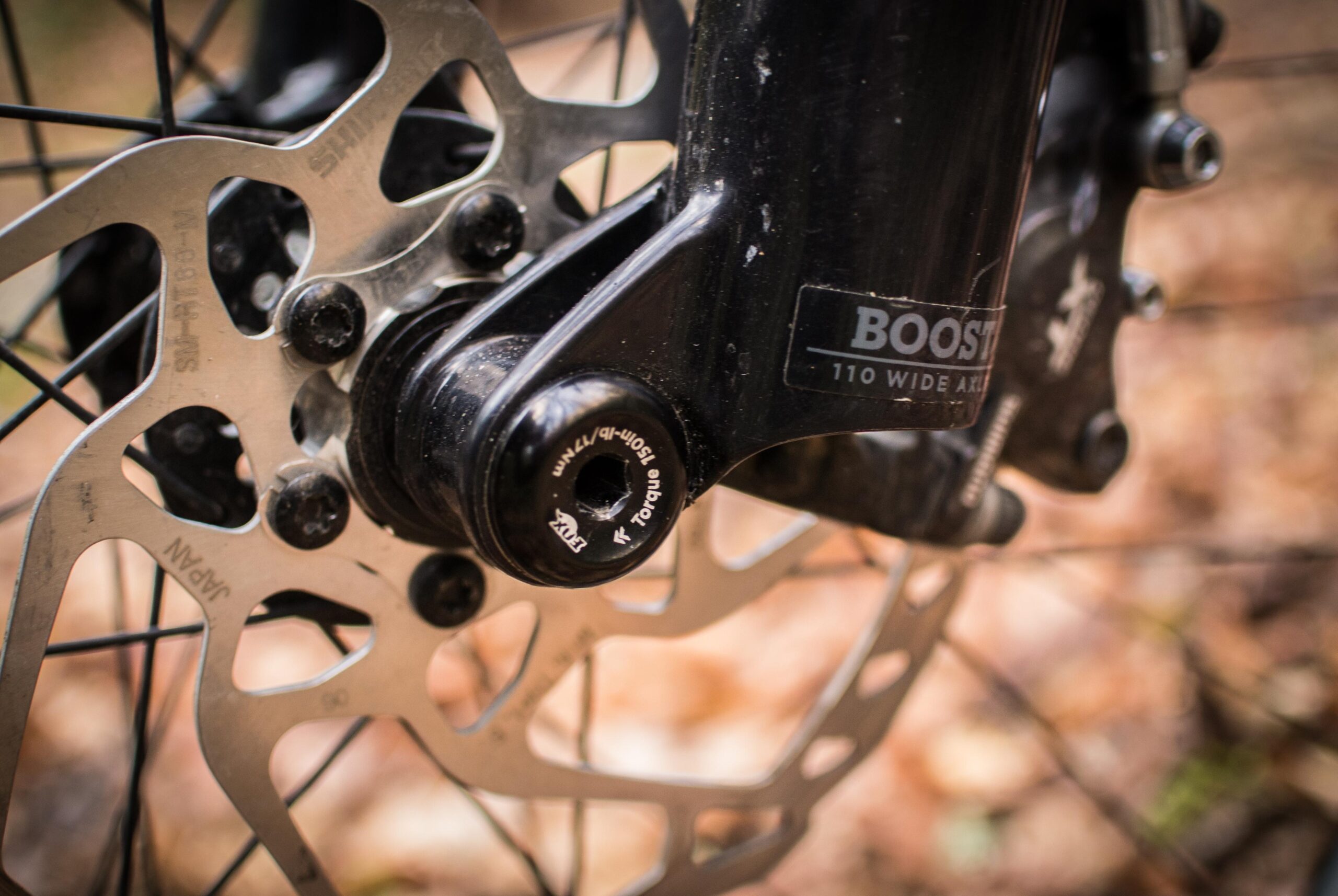Rocky Mountain Element: Close-up view of a mountain bike front wheel hub, featuring a disc brake rotor, black axle, and a "Boost 110 Wide Axle" label, with blurred autumn leaves in the background.
