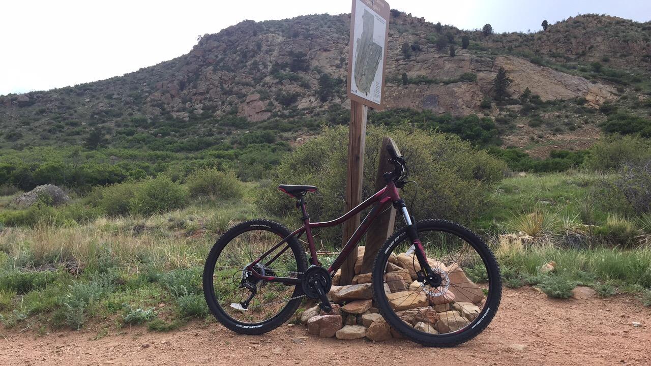 Scott Contessa: A mountain bike leaning against a rock formation on a dirt trail, with a mountainous landscape in the background and a trail sign indicating nearby routes. Lush green vegetation surrounds the area.
