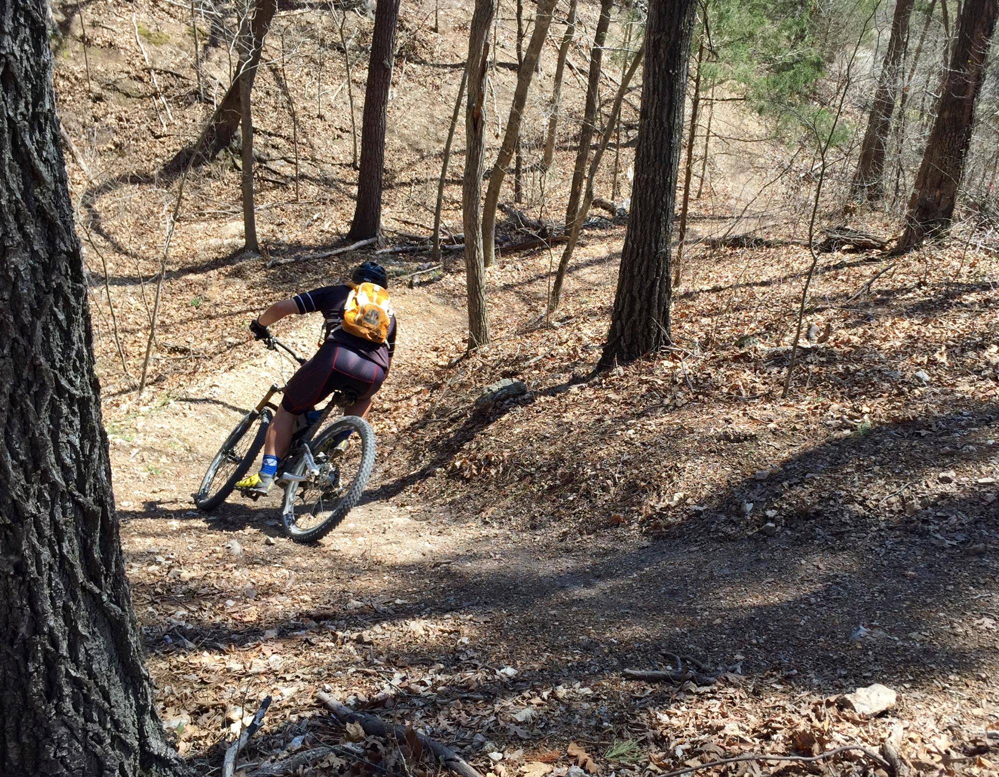 A mountain biker navigating a dirt trail through a wooded area, surrounded by trees and fallen leaves. The cyclist is wearing a helmet and an orange backpack, leaning into a turn on the trail. Back 40 mountain bike trail.