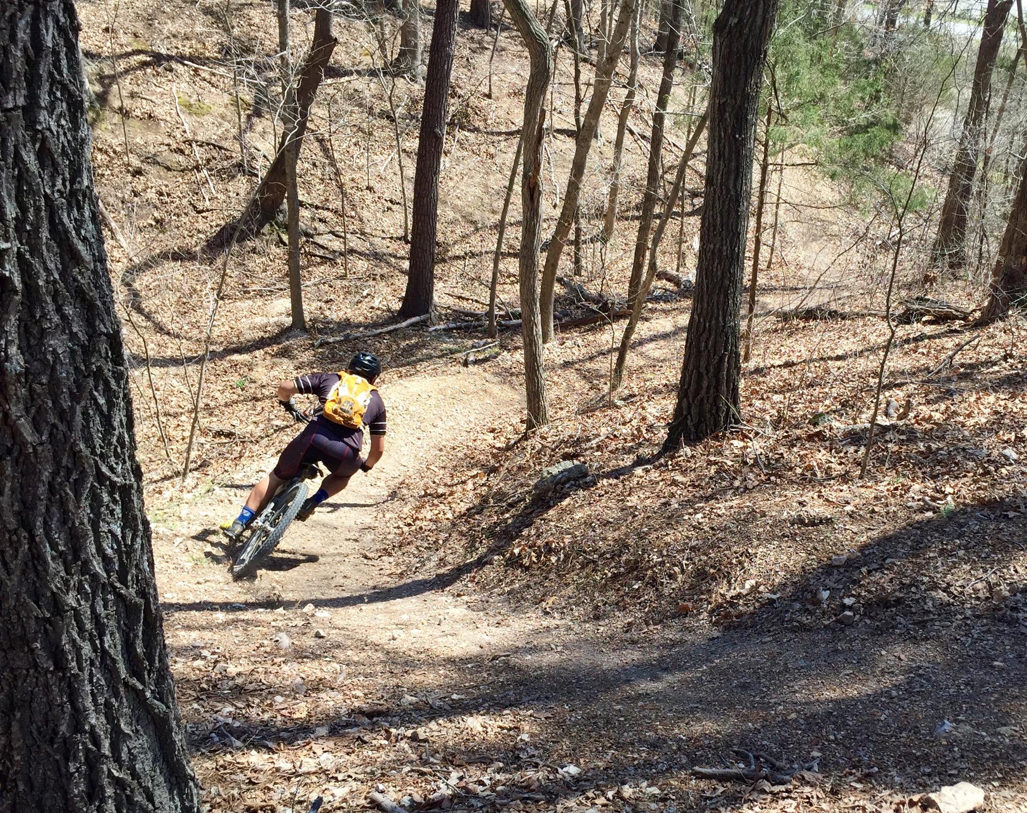 A mountain biker navigates a winding, dirt trail through a wooded area, surrounded by trees and fallen leaves. The cyclist is wearing a helmet and a backpack, leaning into the turn as they ride. Back 40 mountain bike trail.