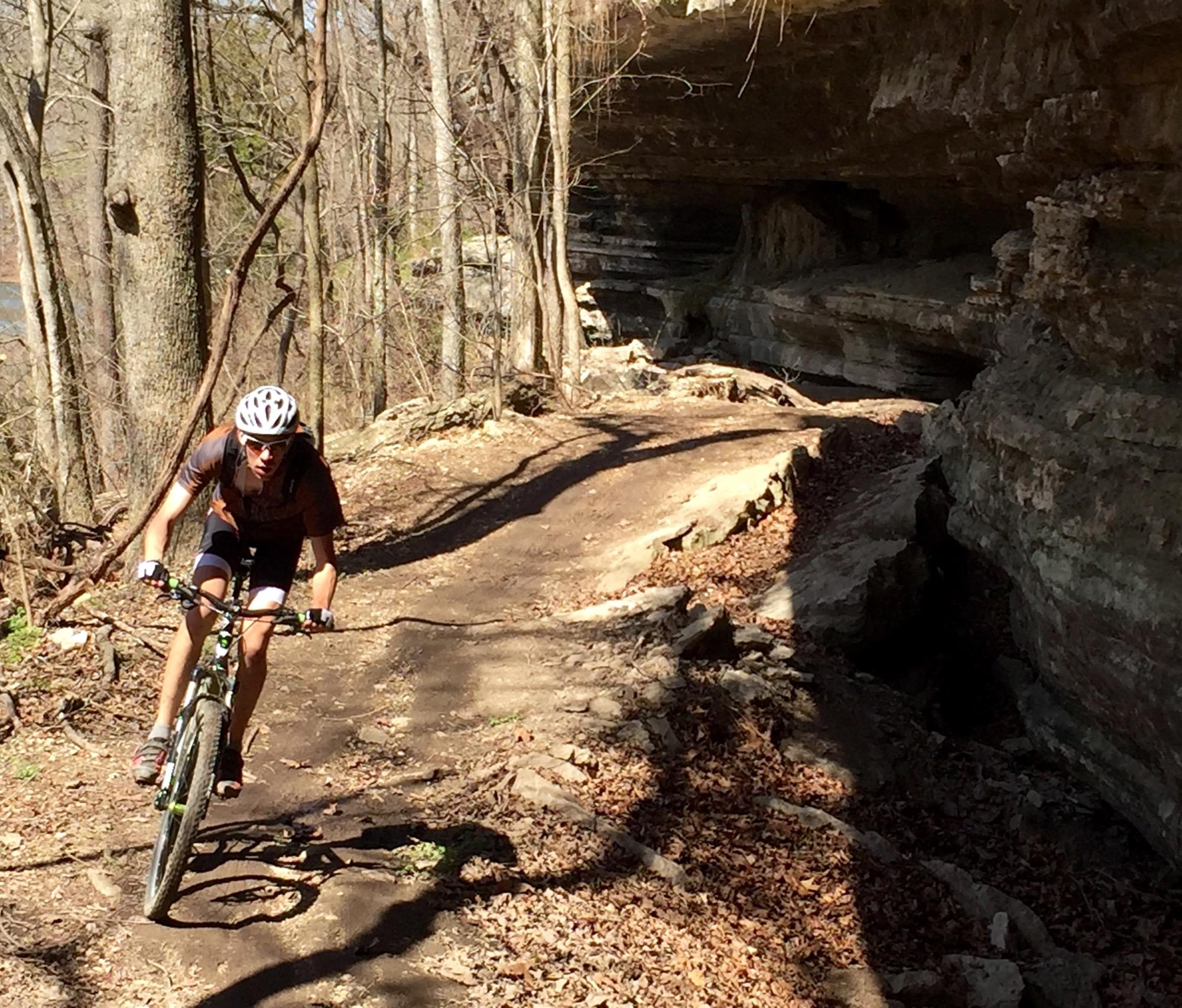 A mountain biker navigates a dirt trail lined with trees and rocky terrain, with a cliff face visible in the background. Sunlight filters through the branches, creating shadows on the path. Back 40 mountain bike trail.