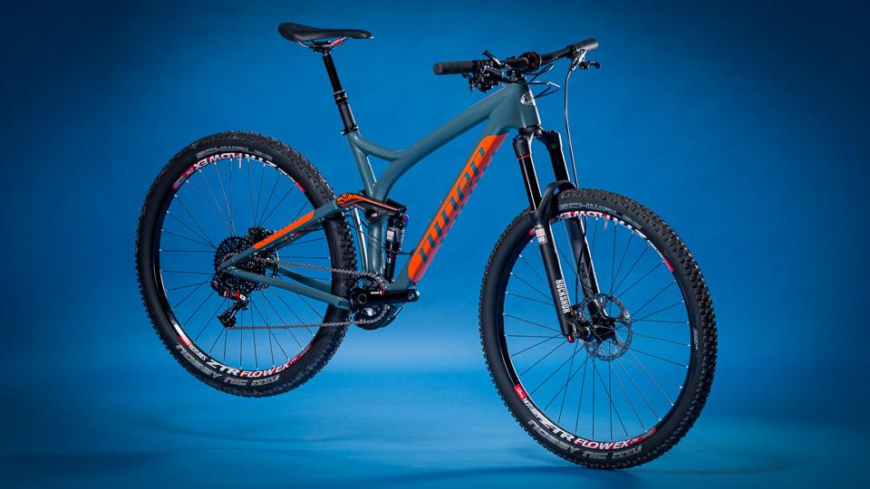 Niner R.I.P. 9 RDO: Mountain bike with a gray frame and orange accents, displayed against a blue background. The bike features large, knobby tires, front suspension forks, and disc brakes, highlighting its design for off-road cycling.