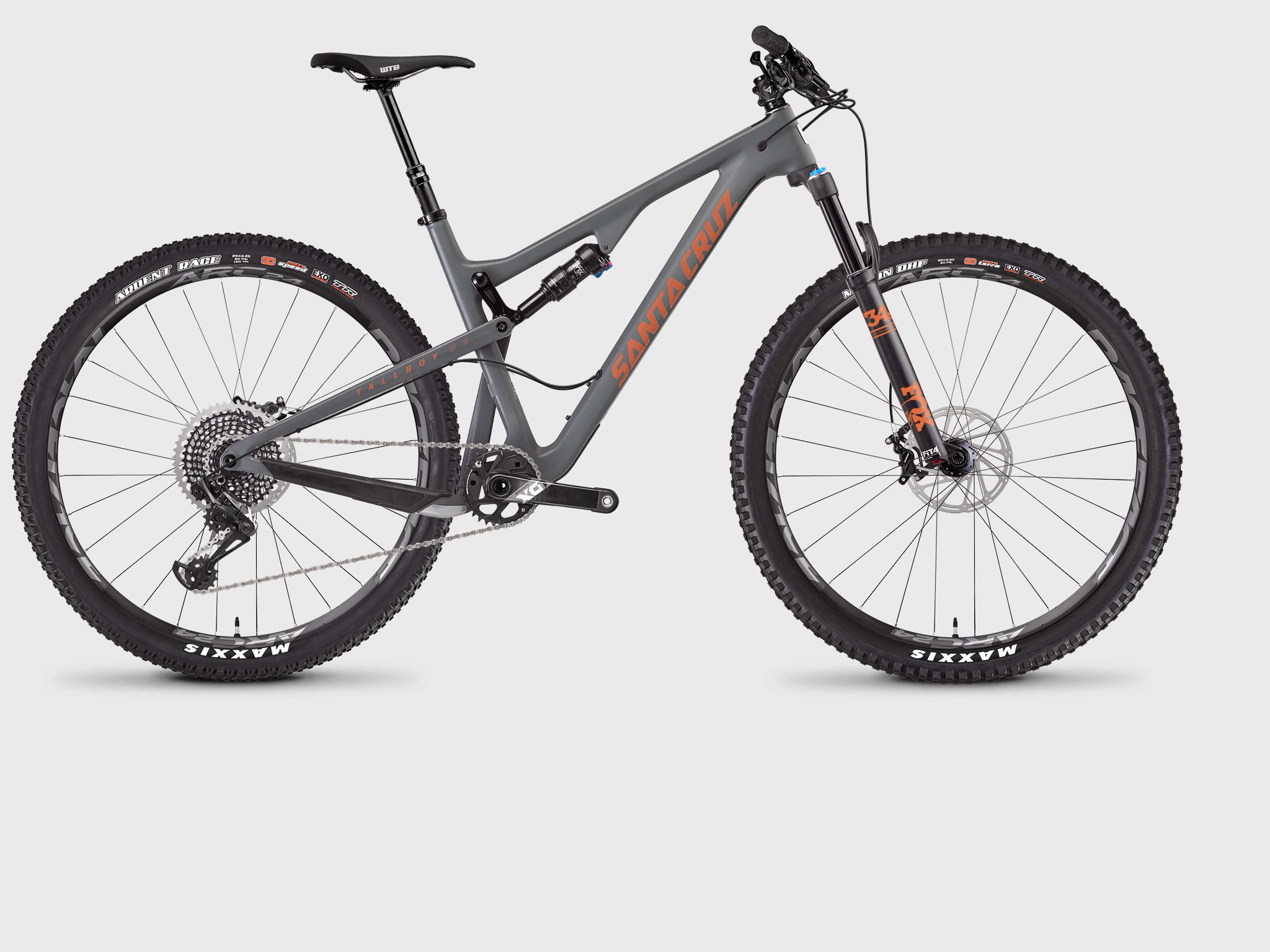 Santa Cruz Tallboy Carbon: A high-performance mountain bike featuring a gray frame, prominently displaying the "Santa Cruz" logo in orange. The bike has wide tires suitable for rough terrain, equipped with a suspension fork and rear shock. The drivetrain is visible, indicating a modern gear setup. The image is set against a neutral background for clarity.