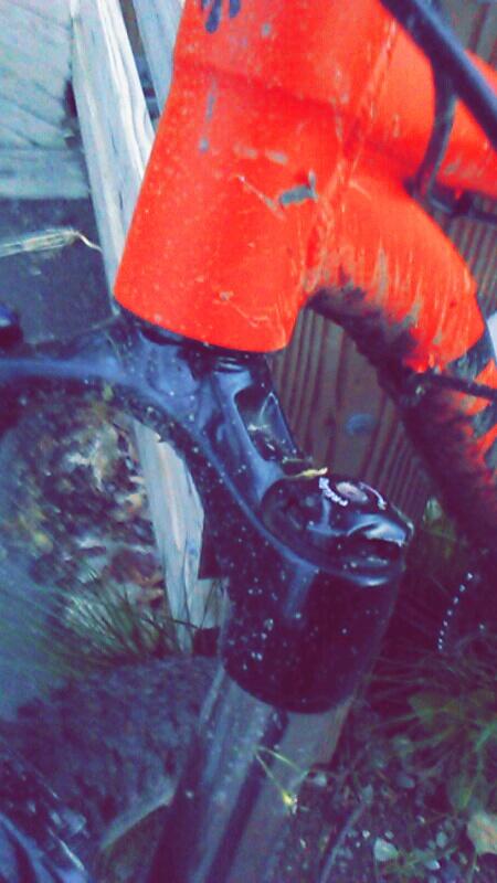 Trek X-Caliber 8: Close-up image of a mountain bike's front fork, showcasing a black suspension fork covered in droplets of water. The bike's frame is partially visible with a bright orange section, indicating it has been used in wet conditions. Background elements include a wooden fence and some greenery, suggesting an outdoor setting.