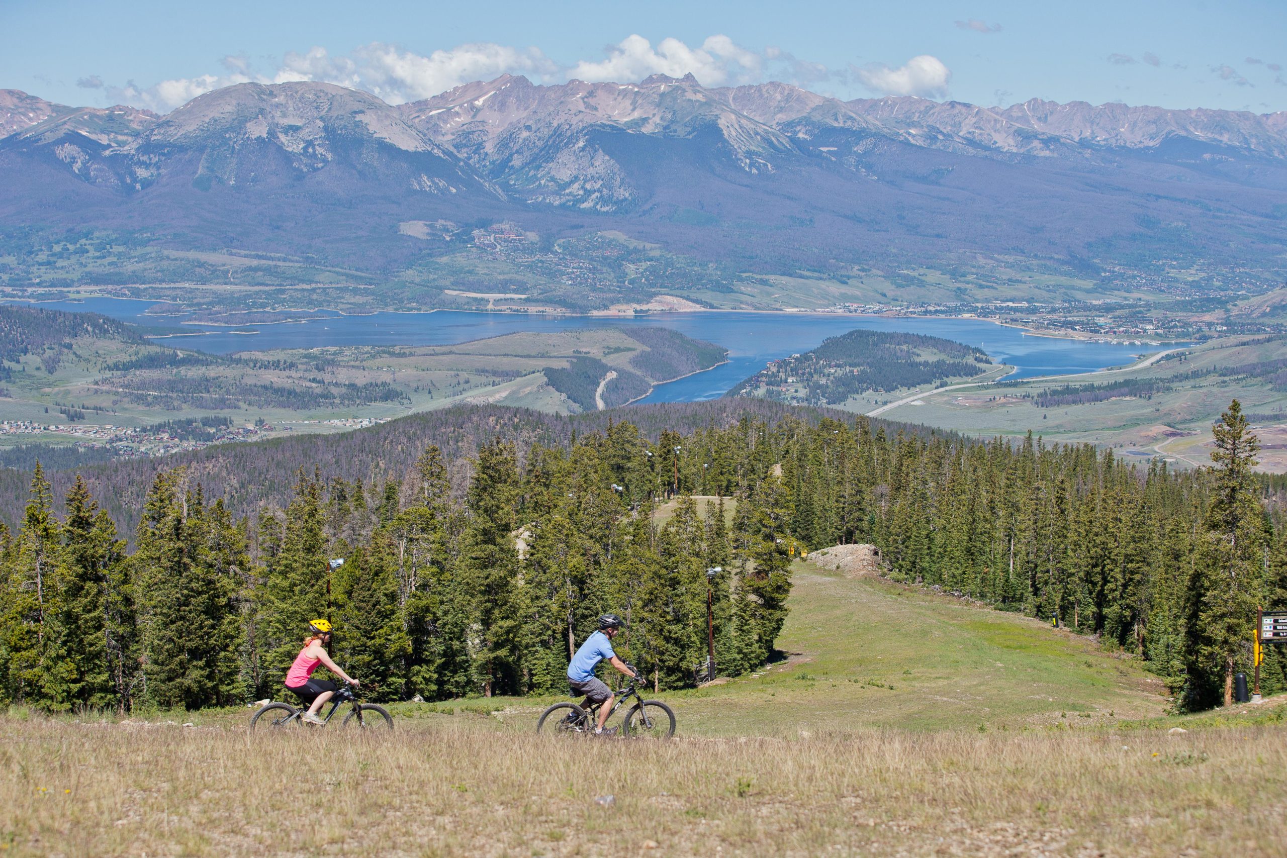 Two mountain bikers ride along a path in a grassy area, surrounded by tall evergreen trees. In the background, a scenic view of mountains and a lake is visible, with a panoramic landscape of valleys and hills under a clear blue sky. Keystone Resort Bike Park mountain bike trail.