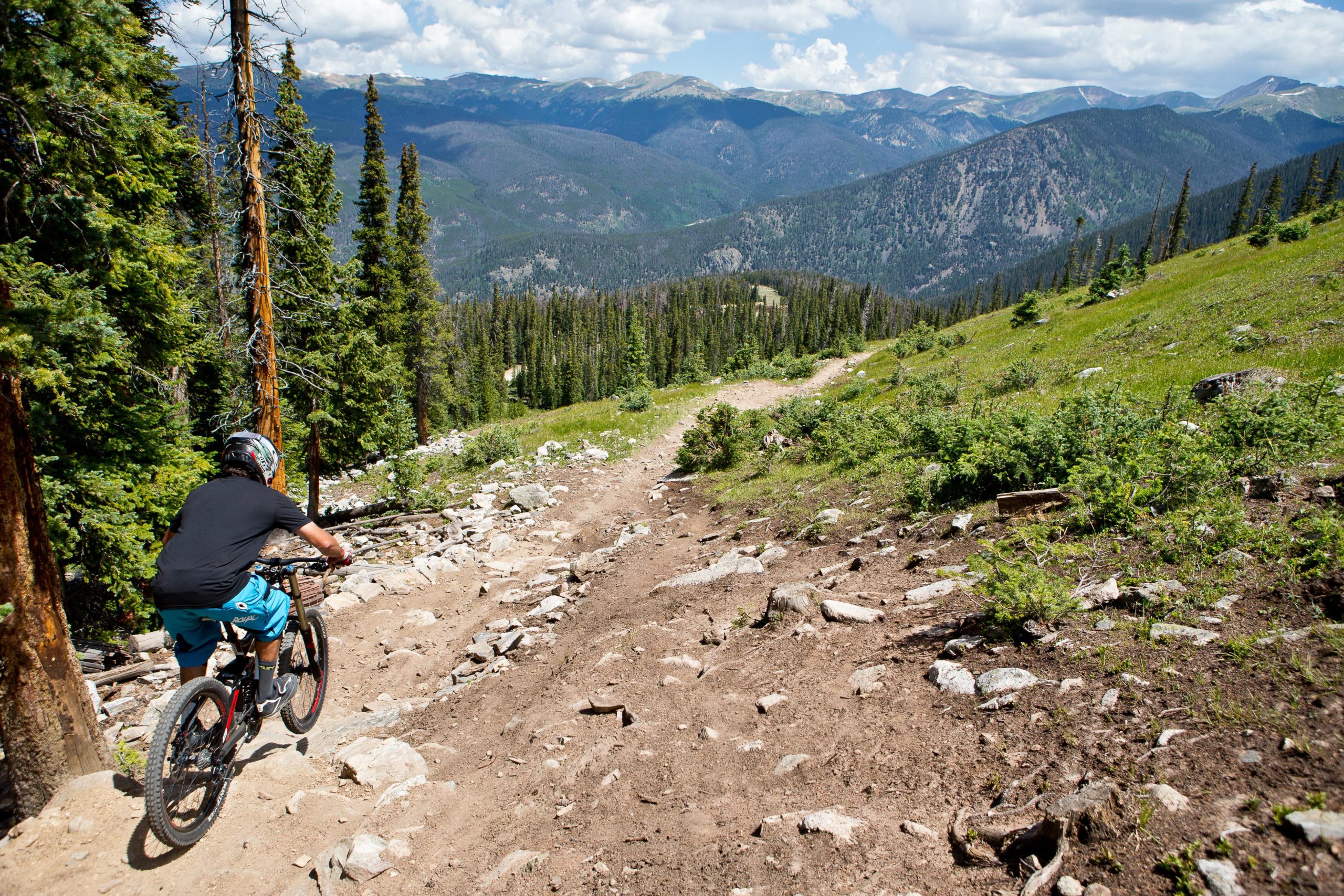 A person riding a mountain bike down a rocky trail surrounded by tall trees and mountains on a sunny day. The scene captures the natural beauty of the outdoors, highlighting the rugged terrain and lush greenery. Keystone Resort Bike Park mountain bike trail.