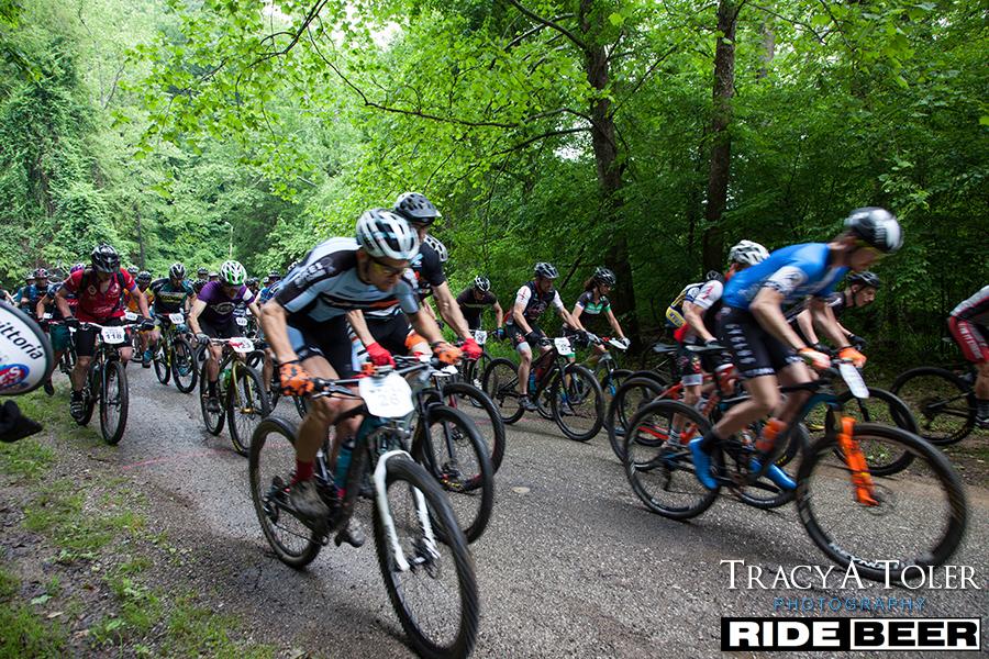 A group of cyclists racing on a dirt path surrounded by lush green trees. The riders are wearing colorful jerseys and helmets, showcasing the dynamic movement and energy of the mountain biking event. Kanawha State Forest Trails mountain bike trail.