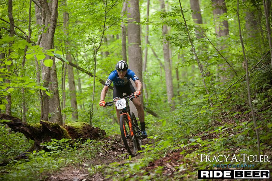 A mountain biker in a blue jersey rides through a lush green forest trail, navigating a dirt path surrounded by tall trees and vibrant foliage. The cyclist, focused and in motion, has a number pinned to his jersey. Sunlight filters through the leaves, highlighting the rich colors of the landscape. Kanawha State Forest Trails mountain bike trail.