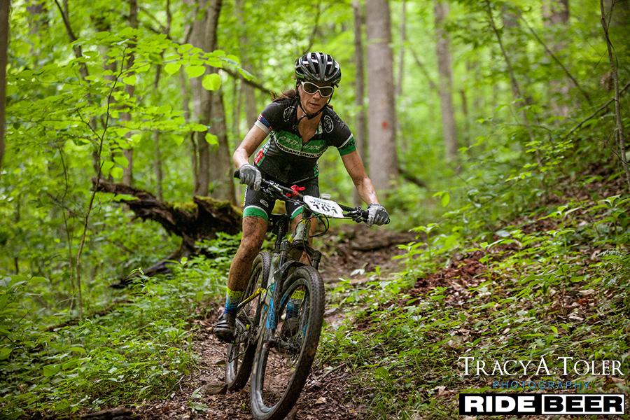 A mountain biker navigates a narrow, wooded trail surrounded by lush greenery, wearing a black helmet and sunglasses. The cyclist, covered in dirt, is focused as they ride on a rugged mountain bike marked with a race number. Kanawha State Forest Trails mountain bike trail.