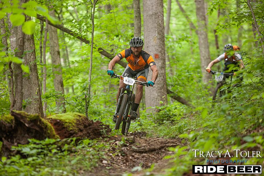 A mountain biker navigates a wooded trail, leaning into a turn while riding through lush greenery. The cyclist is seen wearing a helmet and an orange and gray jersey, with a race number clearly displayed on the front. In the background, another biker can be seen following closely on the trail. Kanawha State Forest Trails mountain bike trail.