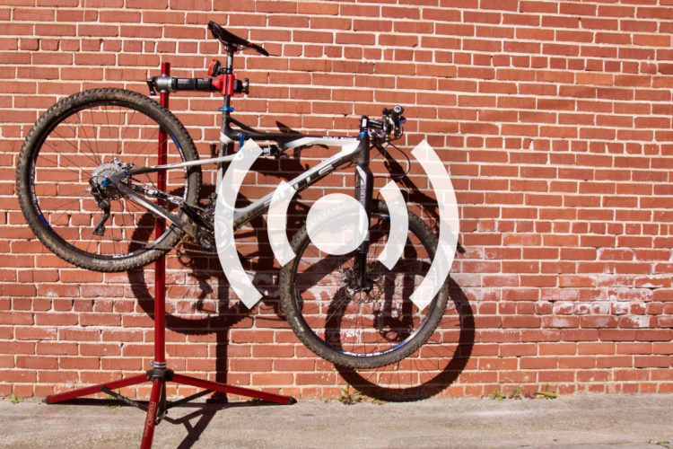 Mountain bike mounted on a repair stand against a brick wall, showcasing its suspension and gears. The bike is slightly dirty, indicating recent use.