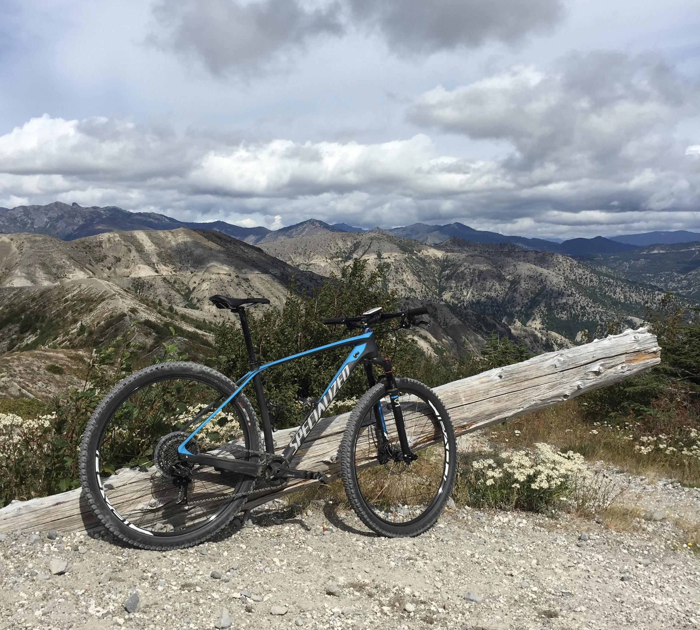 Specialized Stumpjumper FSR Elite: A mountain bike leaning against a log, situated on a rocky trail with expansive mountainous terrain in the background, under a cloudy sky.