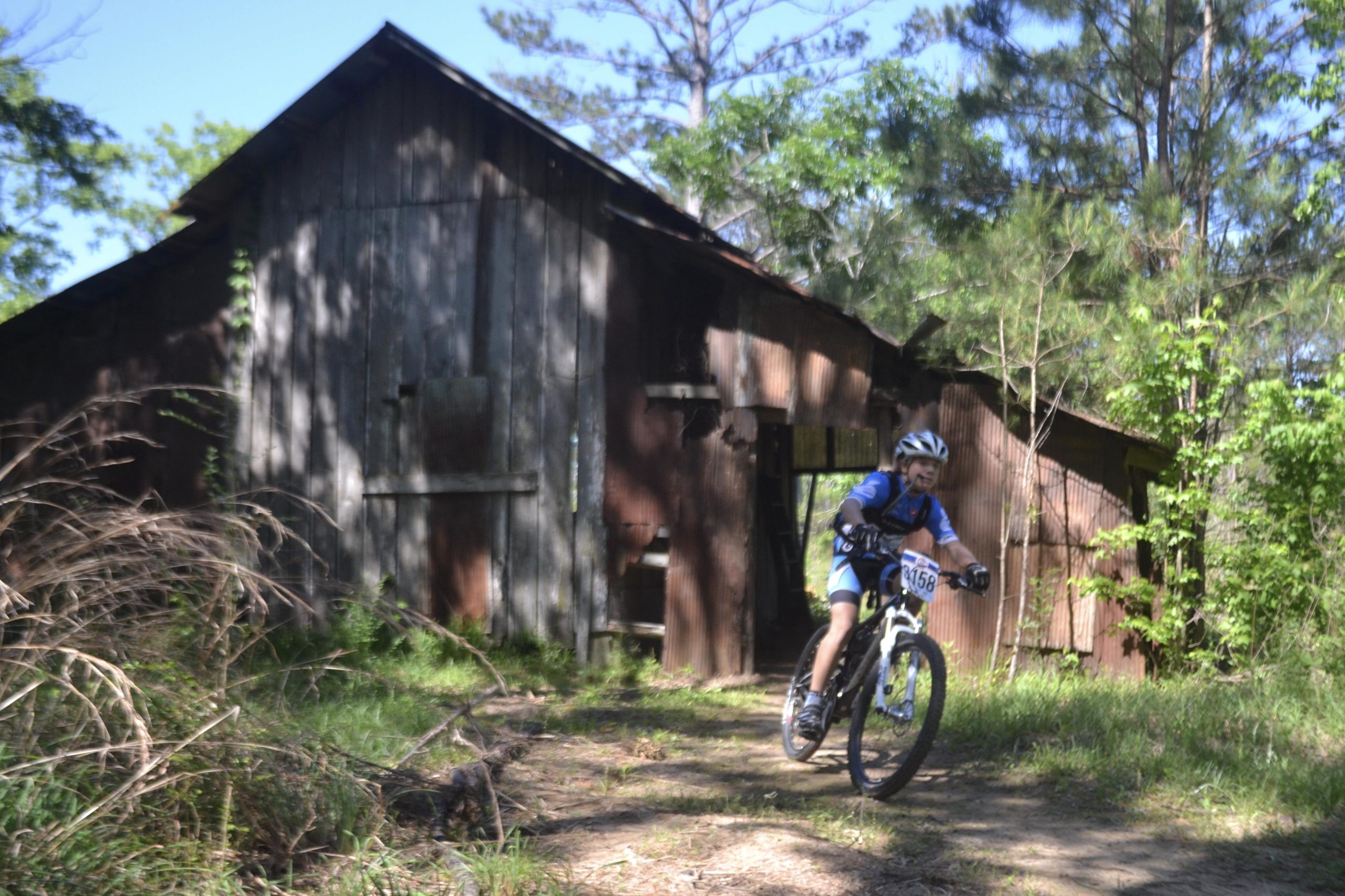 A young cyclist riding a mountain bike along a dirt path with an abandoned wooden building in the background, surrounded by greenery and trees. The cyclist is wearing a helmet and race number, showcasing a dynamic riding pose. Mt. Zion Bike Trails mountain bike trail.