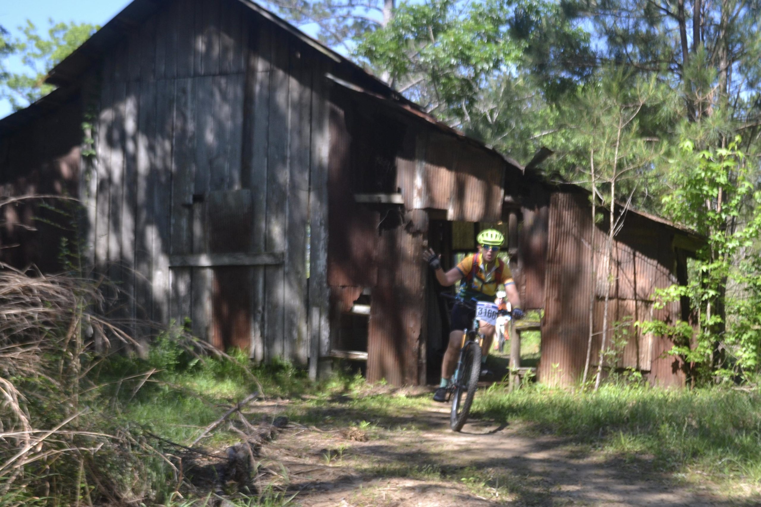 A mountain biker riding on a dirt path near an old, weathered barn surrounded by greenery. The cyclist is wearing a bright helmet and gear, making a hand gesture while navigating the trail. Mt. Zion Bike Trails mountain bike trail.