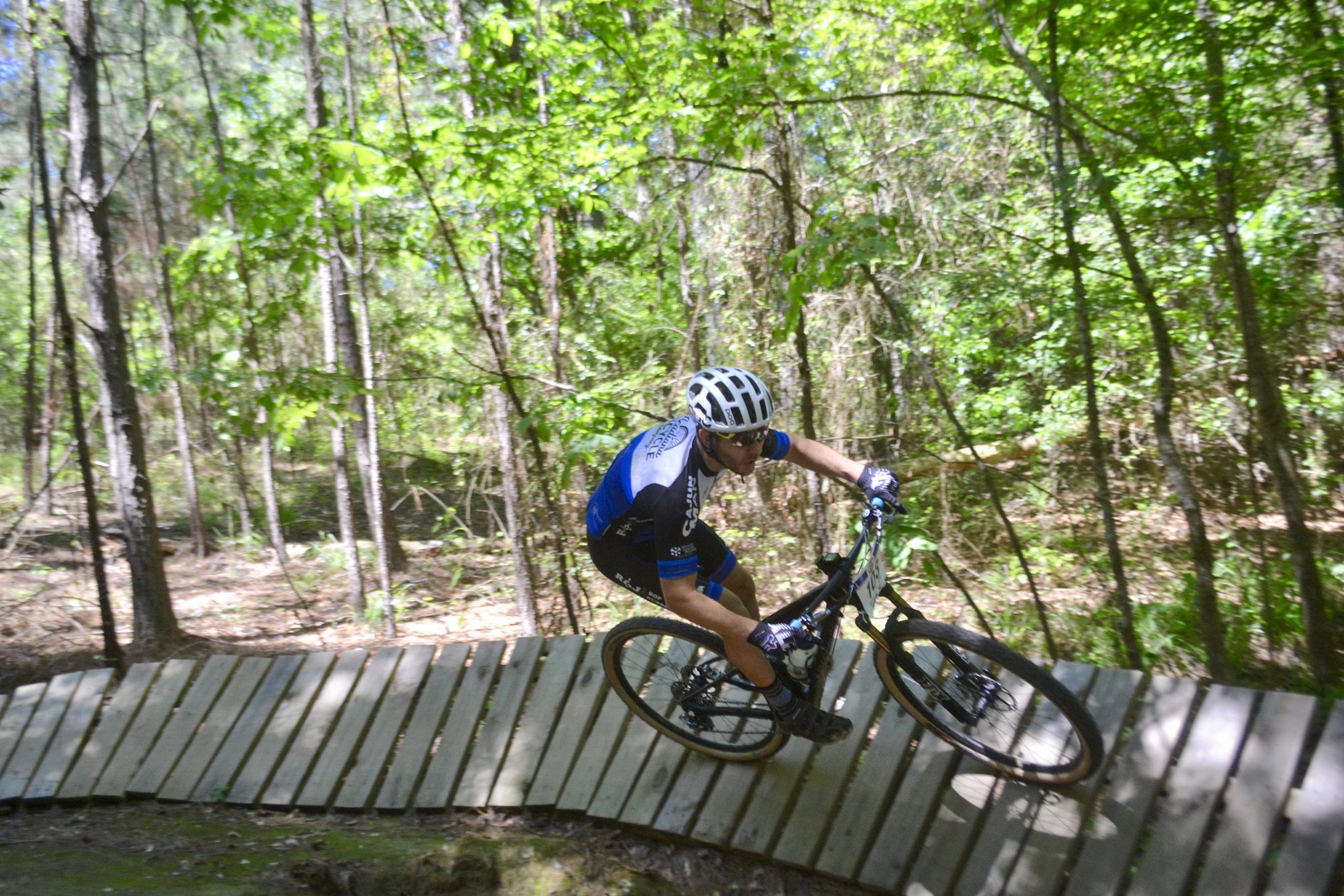 A cyclist in a blue and black jersey rides a mountain bike along a wooden path in a lush green forest, navigating a sharp turn with trees surrounding the trail. Mt. Zion Bike Trails mountain bike trail.