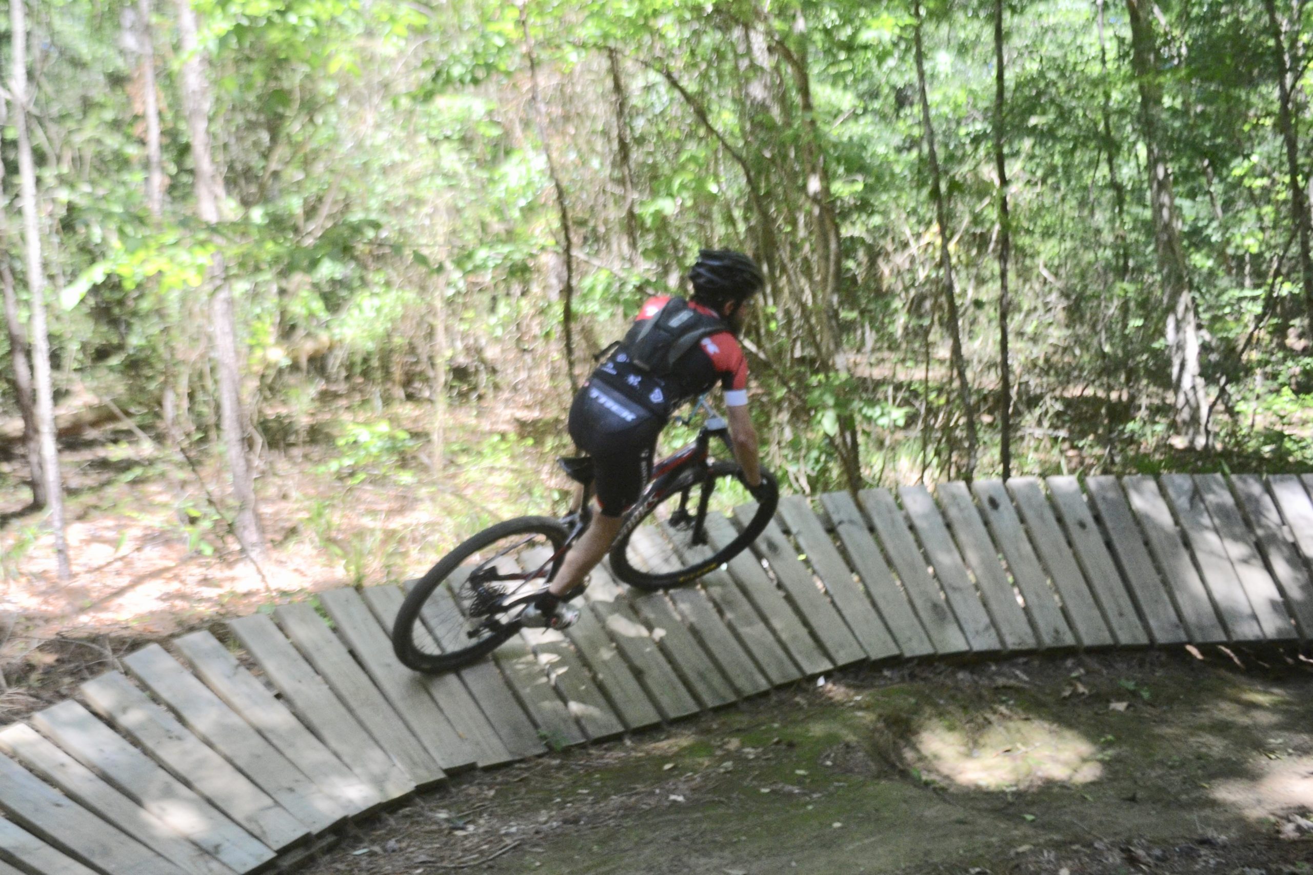 A cyclist navigating a wooden bike path in a forested area, with trees and greenery surrounding the trail. The rider is wearing a helmet and cycling attire, showcasing an action shot as they lean into the curve of the path. Mt. Zion Bike Trails mountain bike trail.