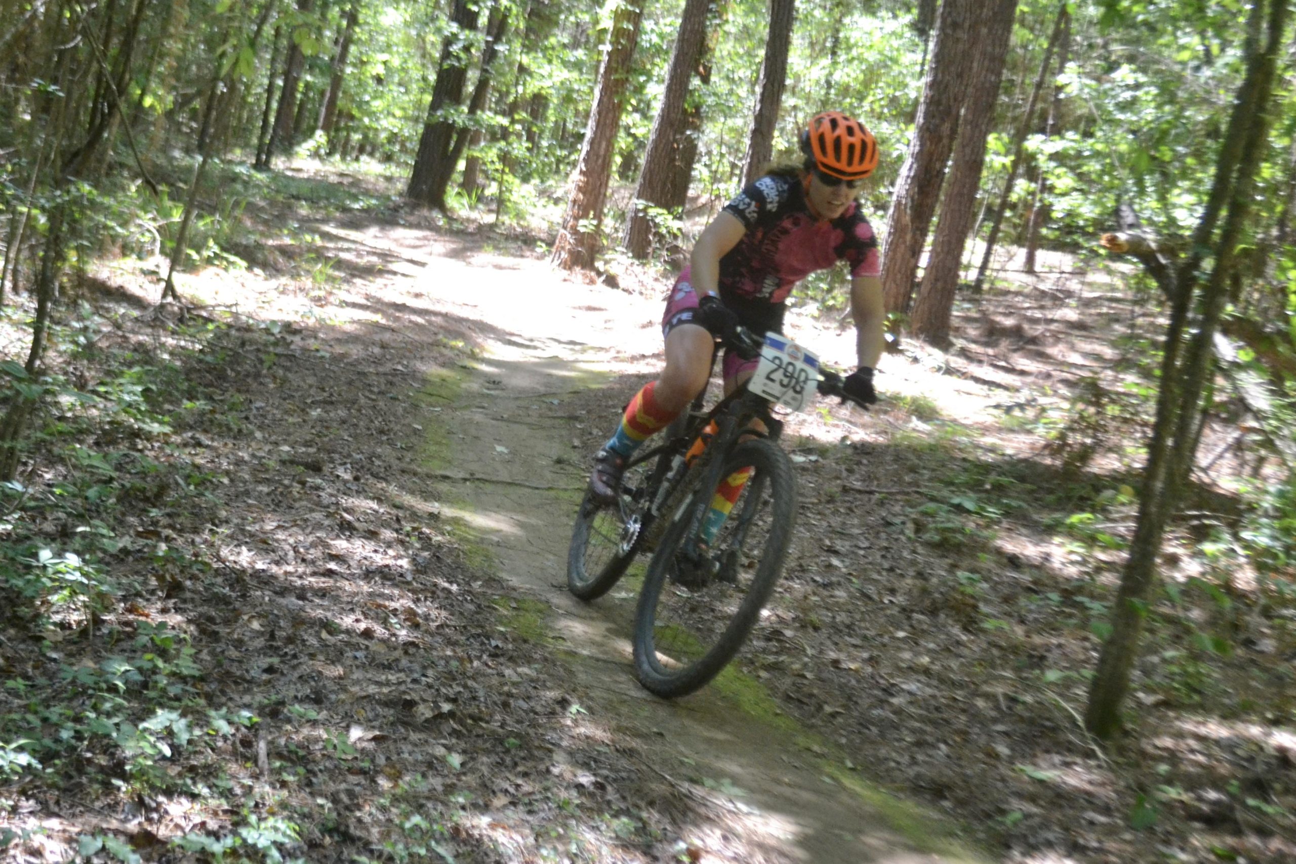 A cyclist riding a mountain bike on a wooded trail, surrounded by trees and leafy ground cover. The rider is wearing a colorful racing outfit with a numbered bib, an orange helmet, and brightly patterned socks, showcasing an energetic and focused posture as they navigate the terrain. Mt. Zion Bike Trails mountain bike trail.