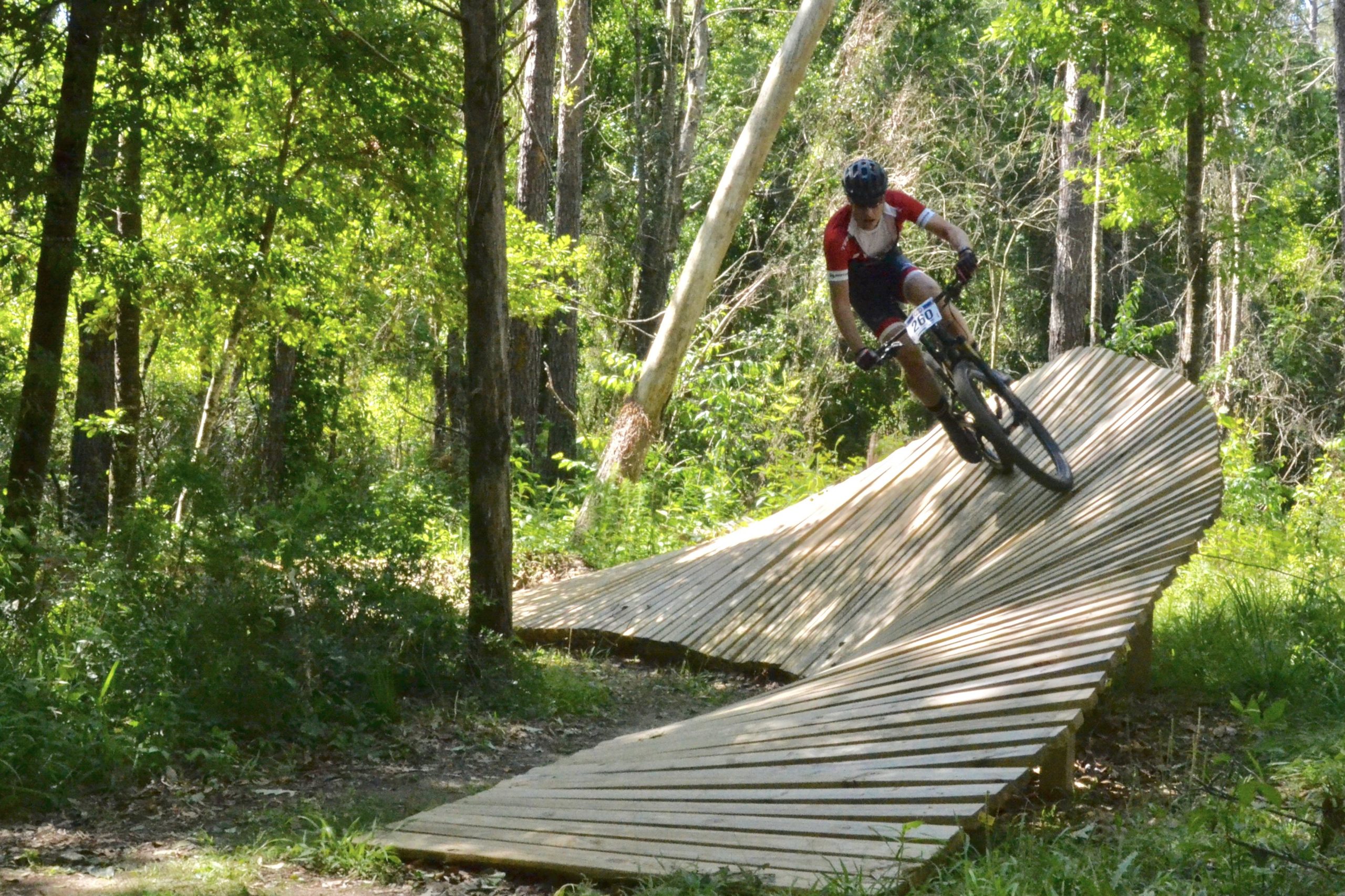 A mountain biker navigating a wooden pump track in a lush green forest, showcasing dynamic movement as they lean into a curve on the track. Sunlight filters through the trees, highlighting the natural surroundings. Mt. Zion Bike Trails mountain bike trail.