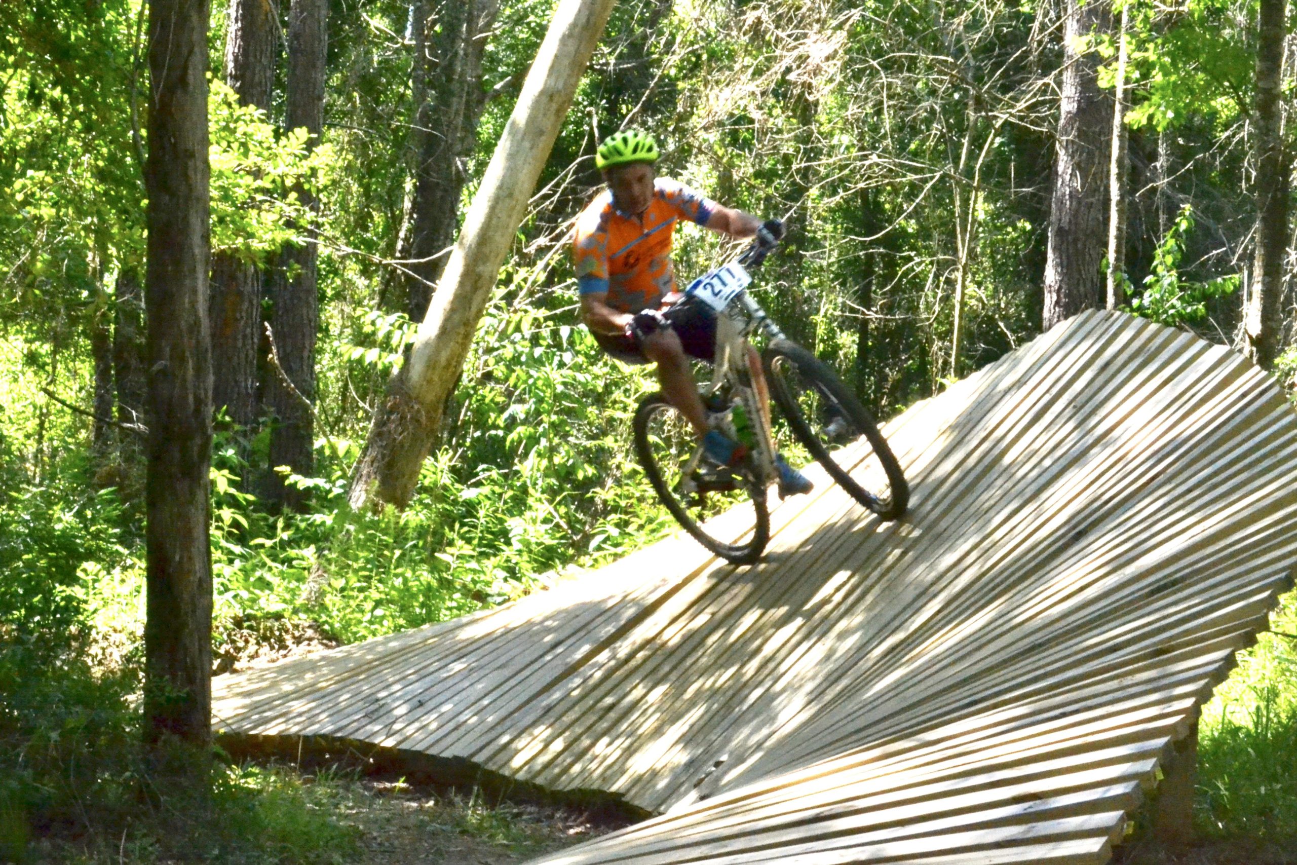 A mountain biker navigating a wooden, undulating trail in a lush green forest. The cyclist, wearing a bright helmet and colorful jersey, is captured mid-motion as they descend the sloped pathway, surrounded by trees and greenery. Mt. Zion Bike Trails mountain bike trail.