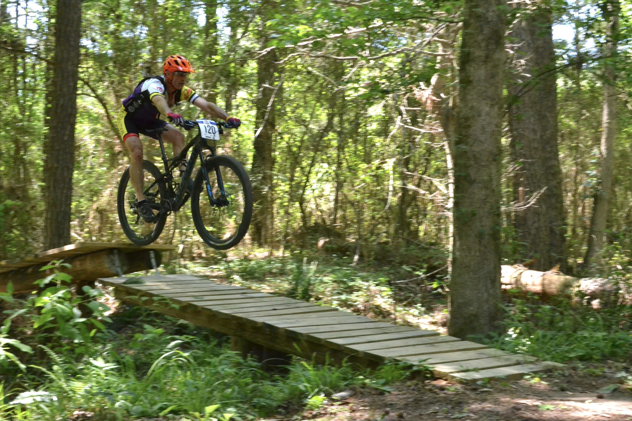 A mountain biker wearing an orange helmet and racing jersey jumps off a wooden bridge in a forested area, showcasing a dynamic action shot amidst greenery and trees. Mt. Zion Bike Trails mountain bike trail.