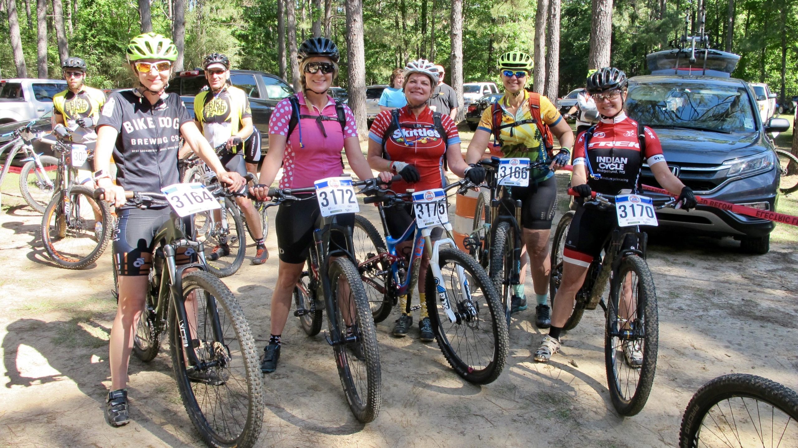 A group of five cyclists, all wearing colorful bike jerseys and helmets, stand on a dirt path with mountain bikes. They are smiling and posing for the camera, with race numbers visibly attached to their bikes. In the background, trees and parked cars can be seen, indicating a biking event or competition setting. Mt. Zion Bike Trails mountain bike trail.