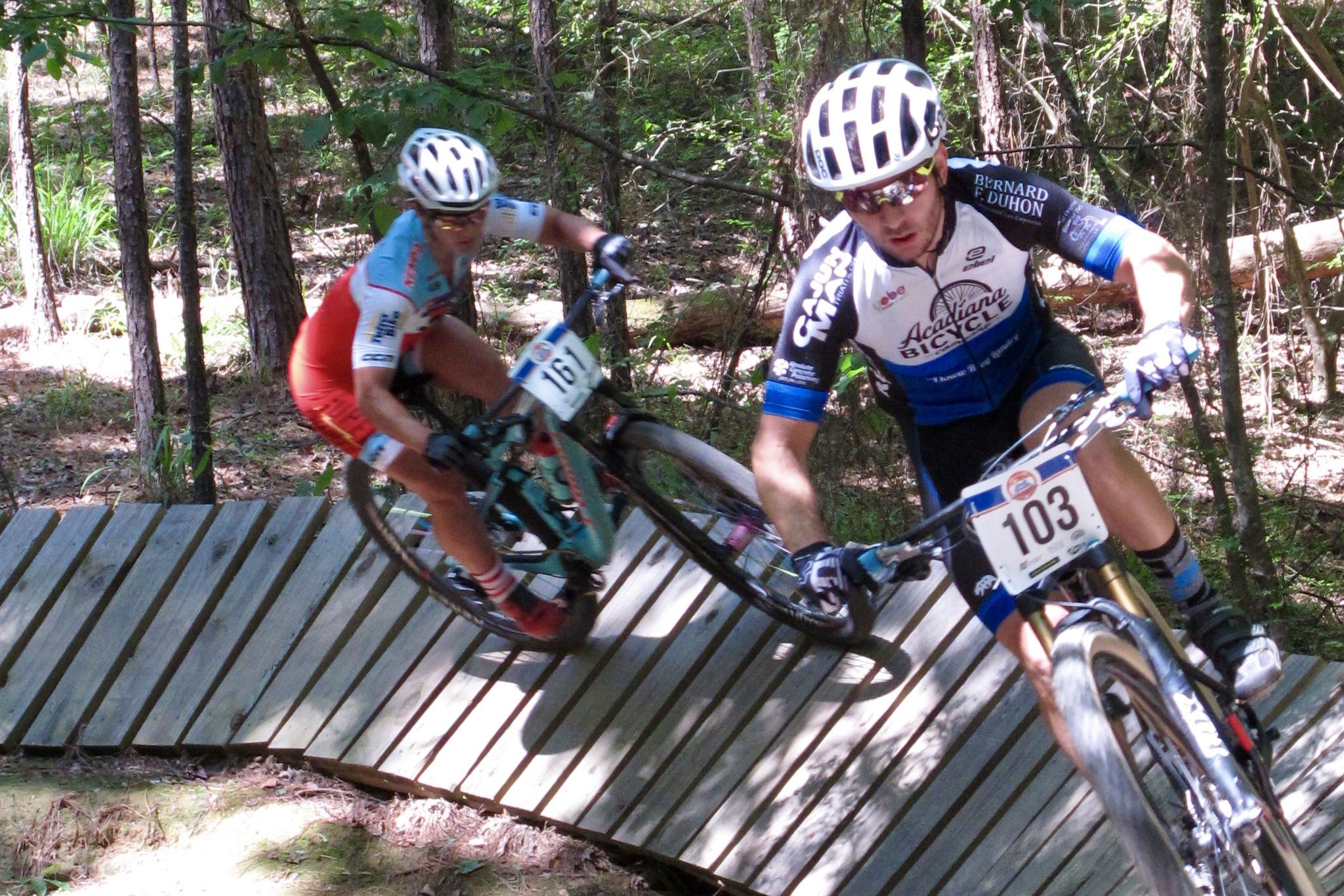 Two mountain bikers navigate a wooden boardwalk trail in a forested area. One rider, prominent in the foreground, is wearing a blue and white jersey with the number 103, while the other, in the background, is dressed in a red and orange jersey with the number 161. Sunlight filters through the trees, highlighting the dynamic movement as they lean into a turn. Mt. Zion Bike Trails mountain bike trail.