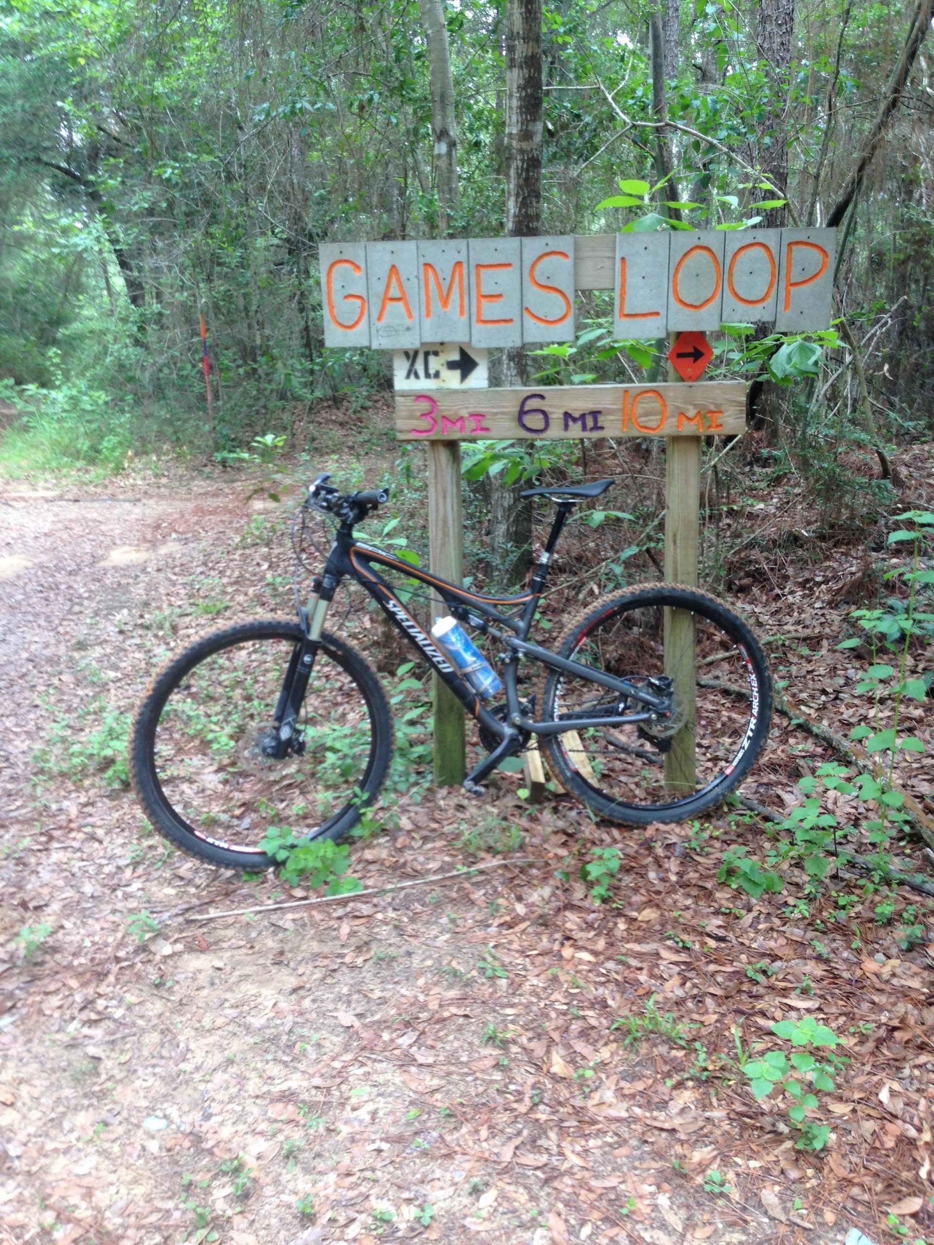 A mountain bike leaning against a wooden sign that reads "GAMES LOOP" with arrows indicating directions. The sign also displays distances for trails: 3 miles, 6 miles, and 10 miles. The surrounding area is lush with trees and leafy undergrowth, indicating a natural outdoor setting. UWF Mountain Bike Trails mountain bike trail.