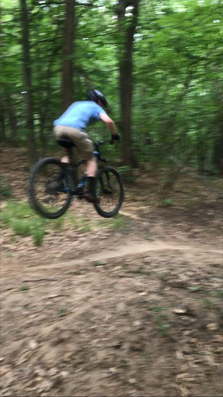 A person riding a mountain bike in a wooded area, captured mid-jump over a dirt path, with trees and foliage blurred in the background, indicating motion and excitement. Merrell Trail mountain bike trail.
